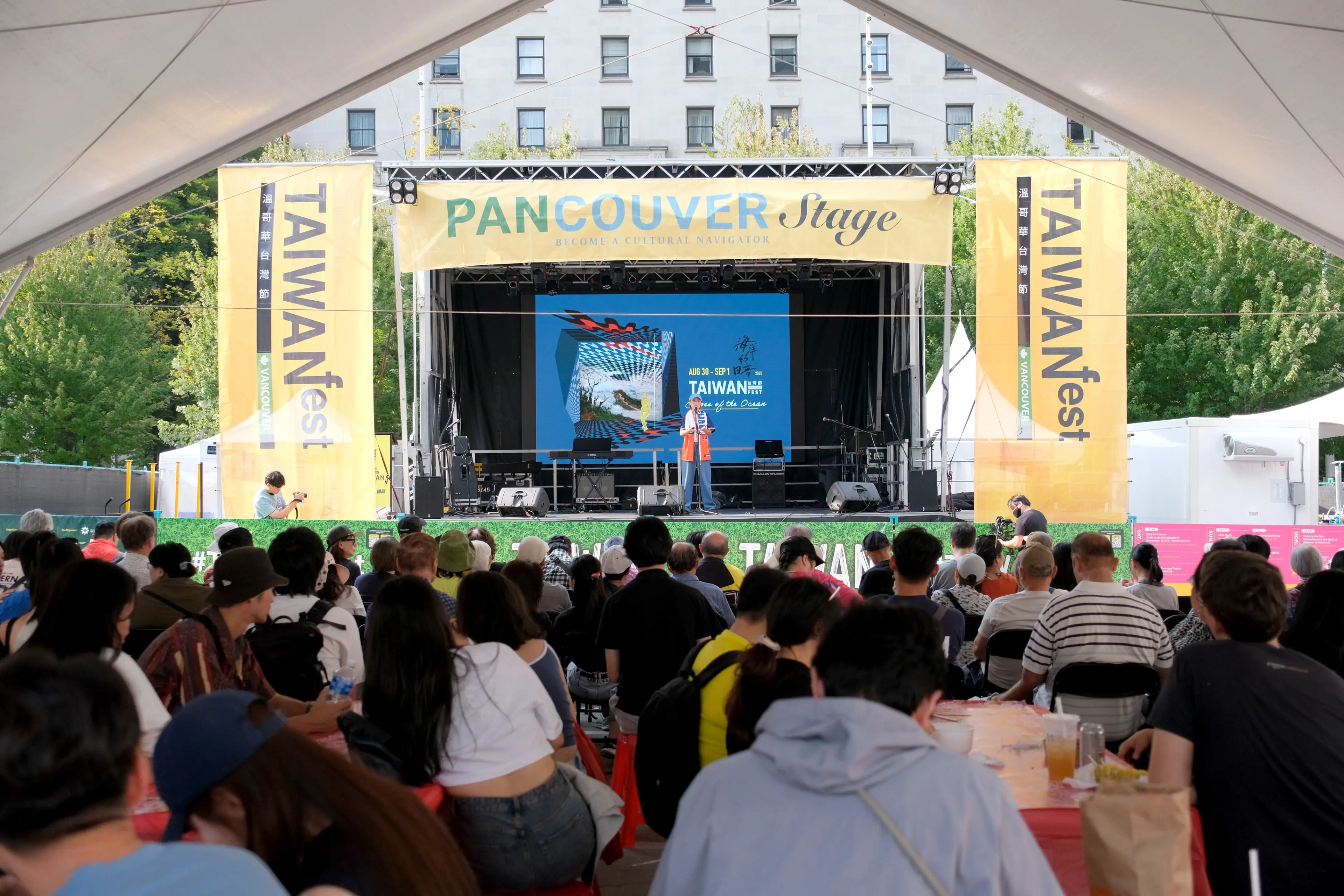 Crowd seated under a tent watching a speaker on the PANCOVER Stage at Taiwan Fest with yellow banners on each side.