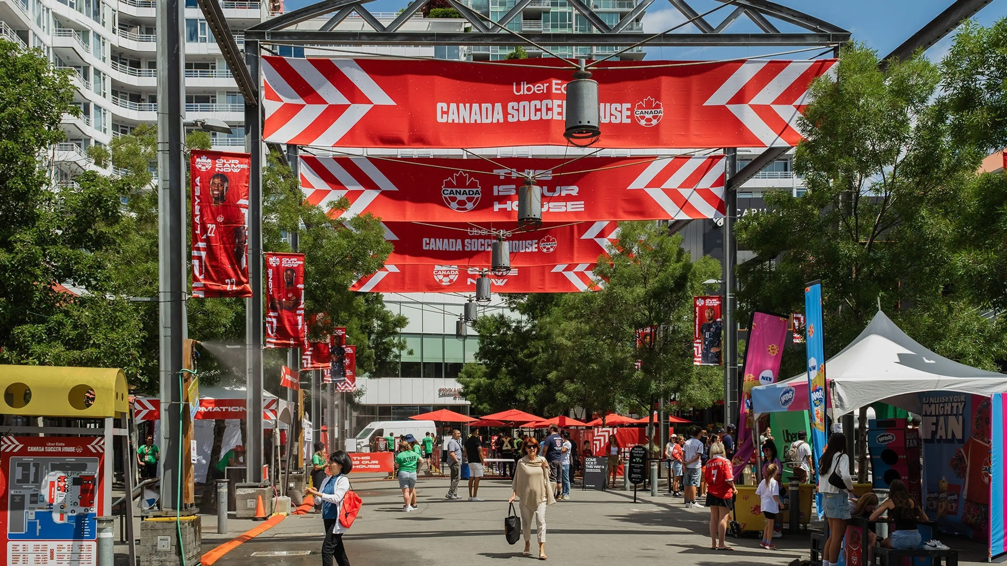 Outdoor event with red Canada Soccer House banners, people walking and standing near booths and tents on a sunny day.