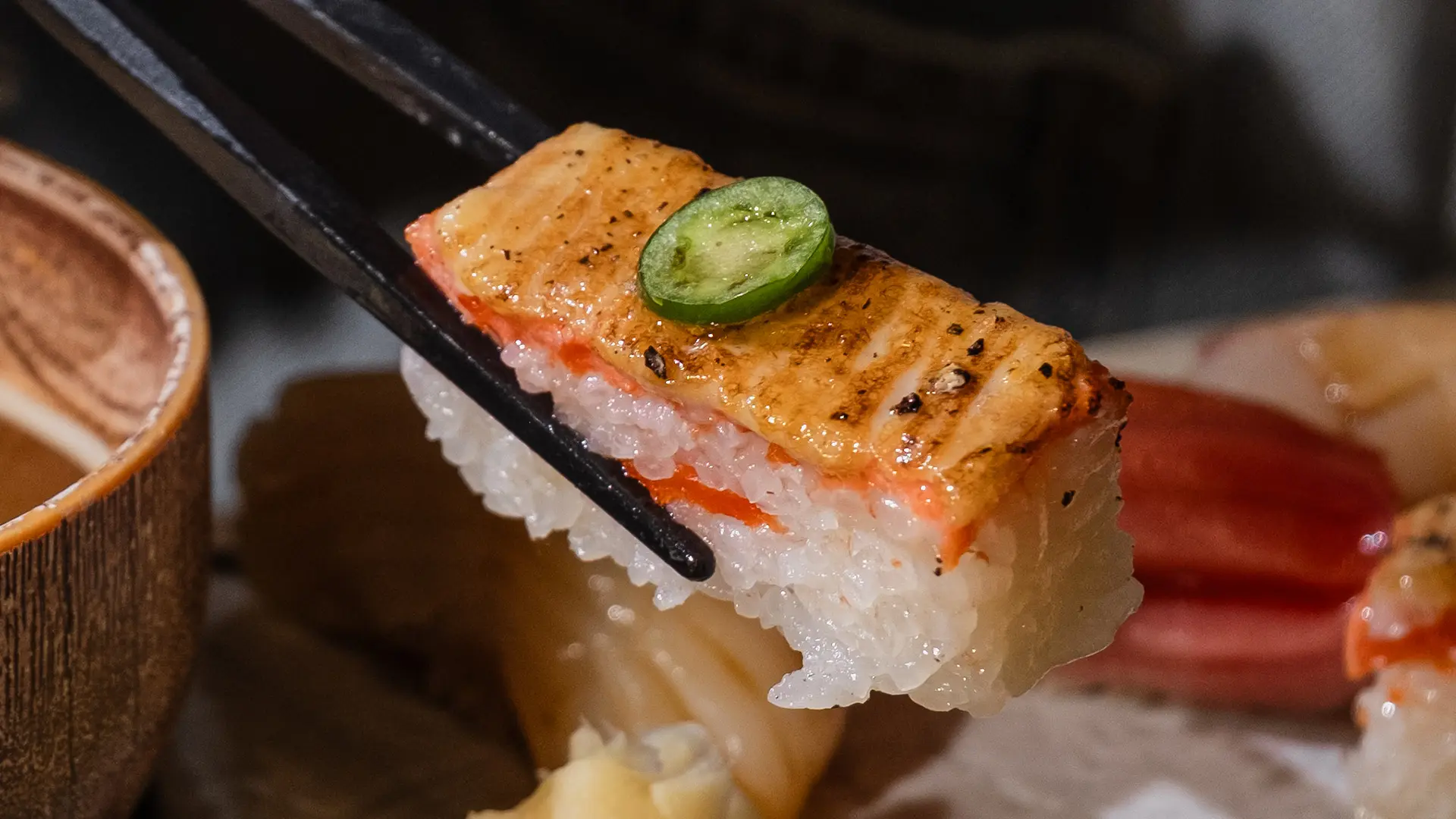 Chopsticks holding a piece of sushi with grilled salmon, rice, and a slice of green chili on top.