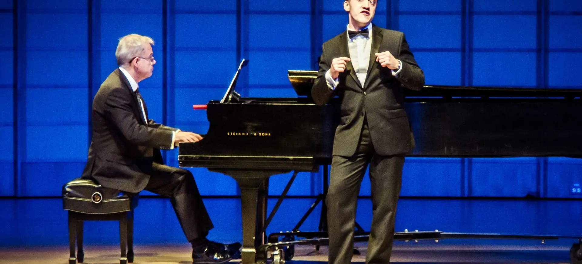 A person in a tuxedo singing on stage with a pianist playing a grand piano in the background.