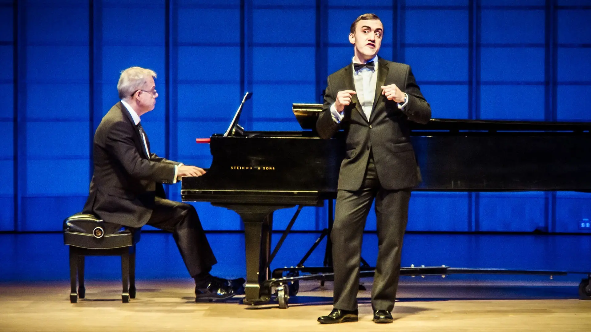 A person in a tuxedo singing on stage with a pianist playing a grand piano in the background.