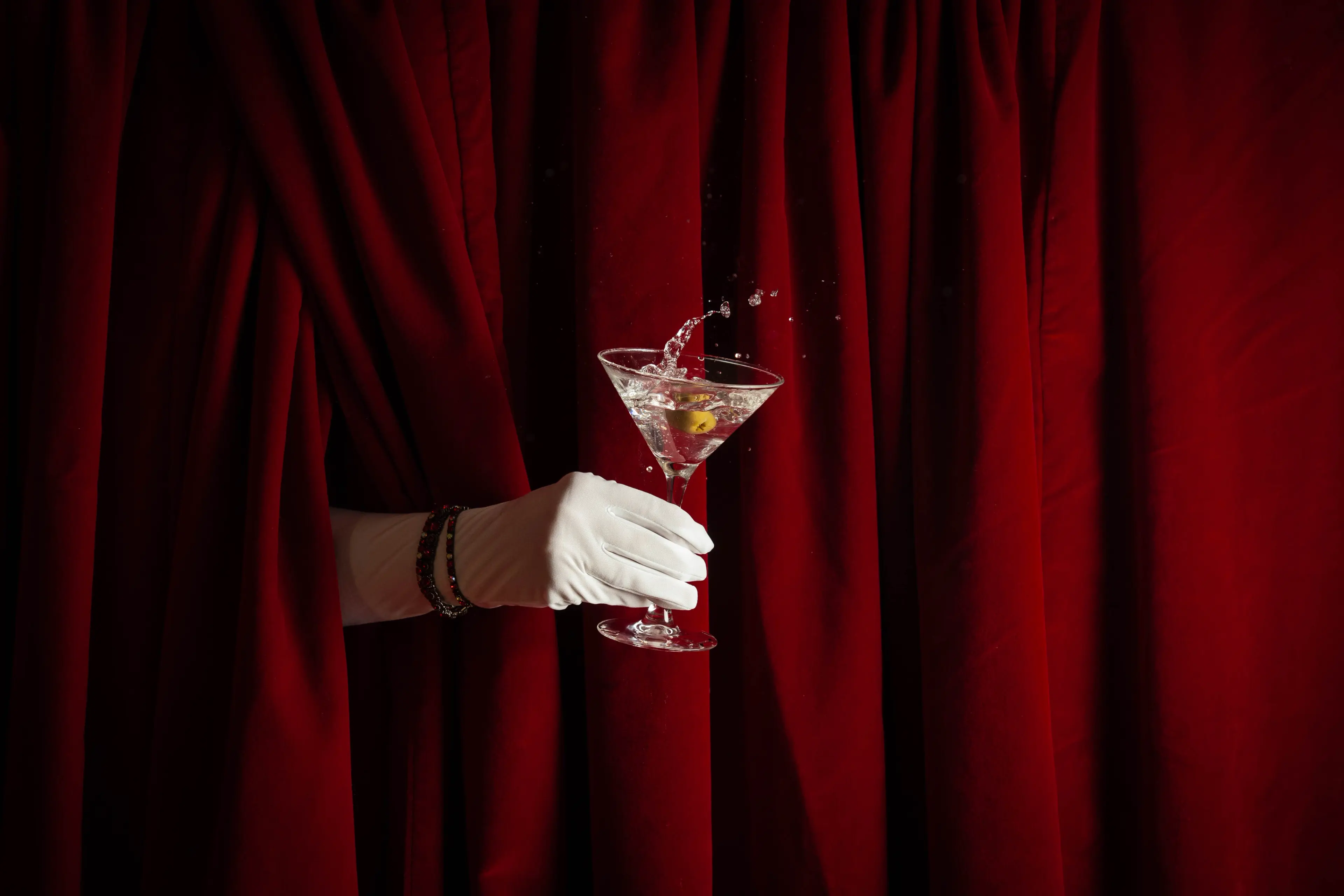 A white gloved hand holding a cocktail glass from behind a red curtain