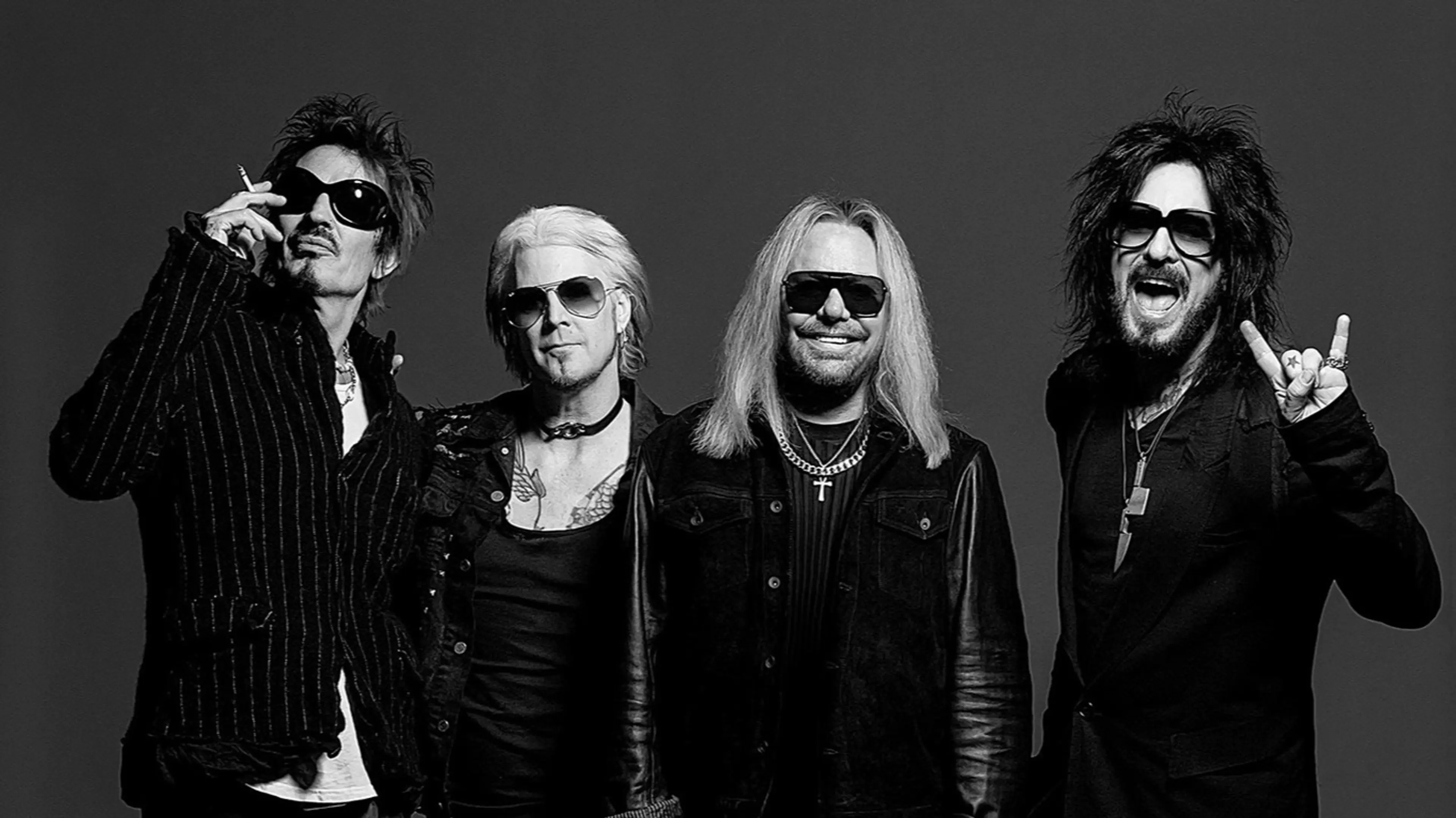 Four people wearing sunglasses and dark clothing posing against a plain dark background, one holding a cigarette and another making a rock hand gesture.