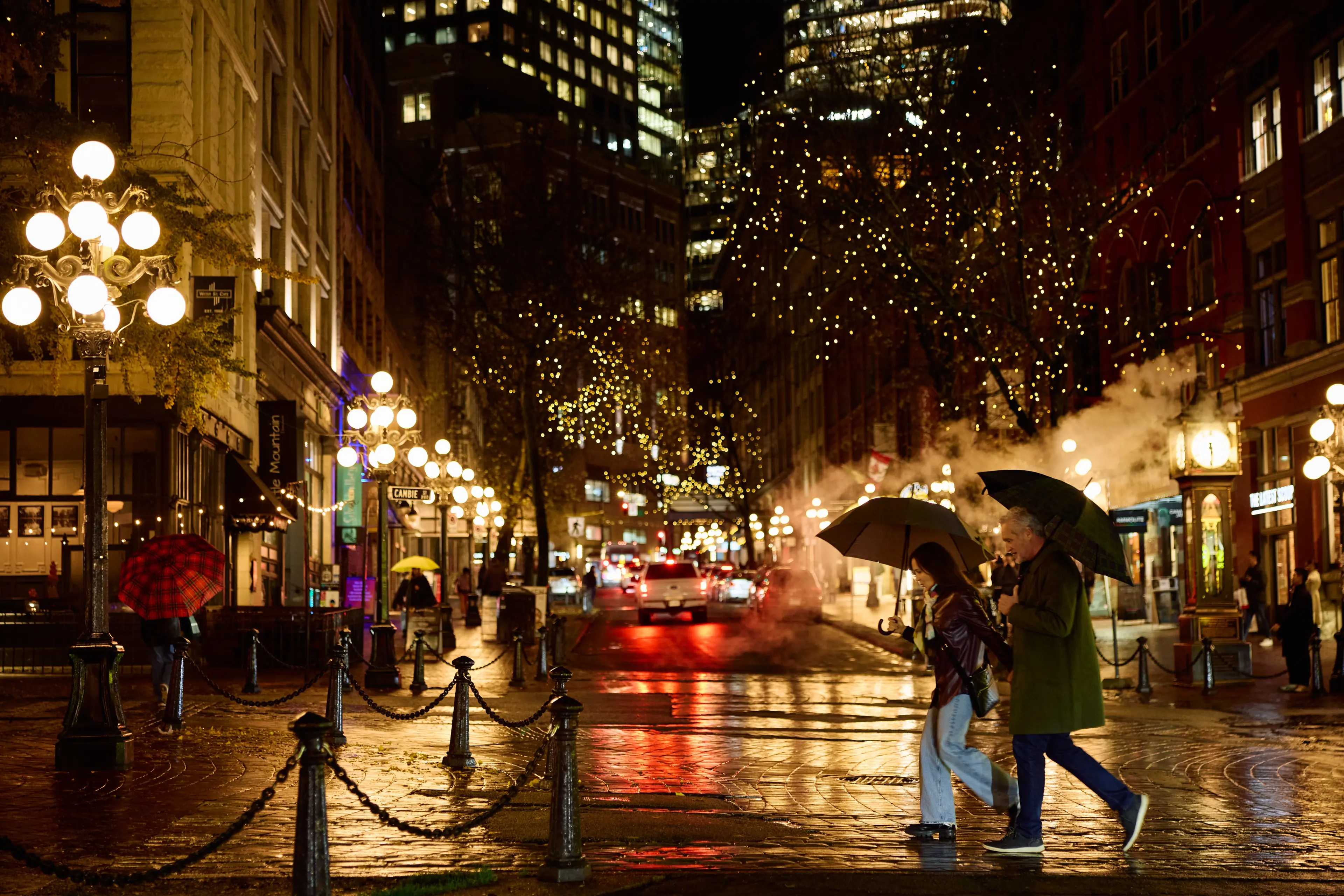 Two people walking with umbrellas on a wet city street at night with illuminated street lamps and buildings.