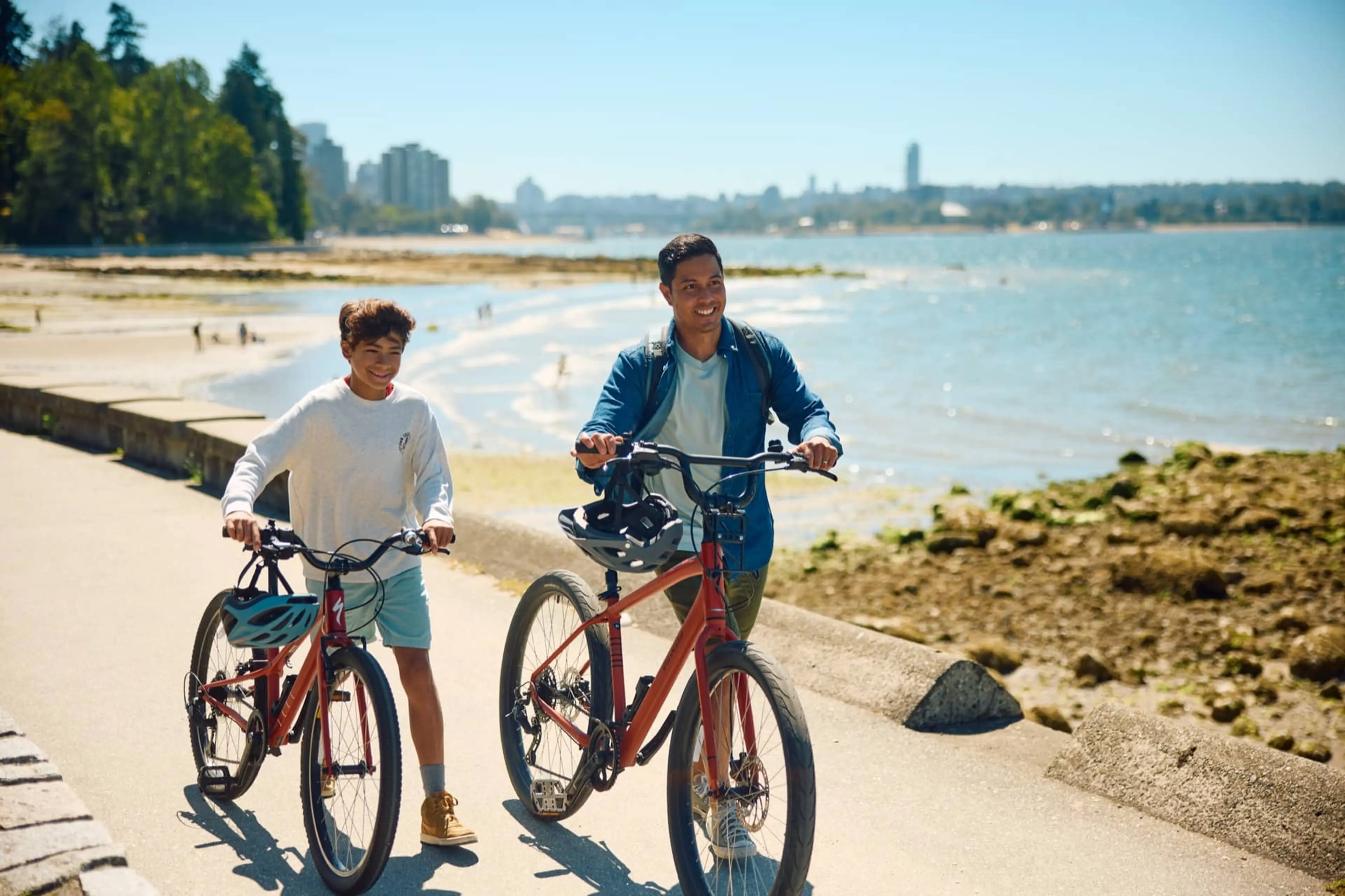 Two people walking with bicycles on a sunny path by the water with city buildings in the background.