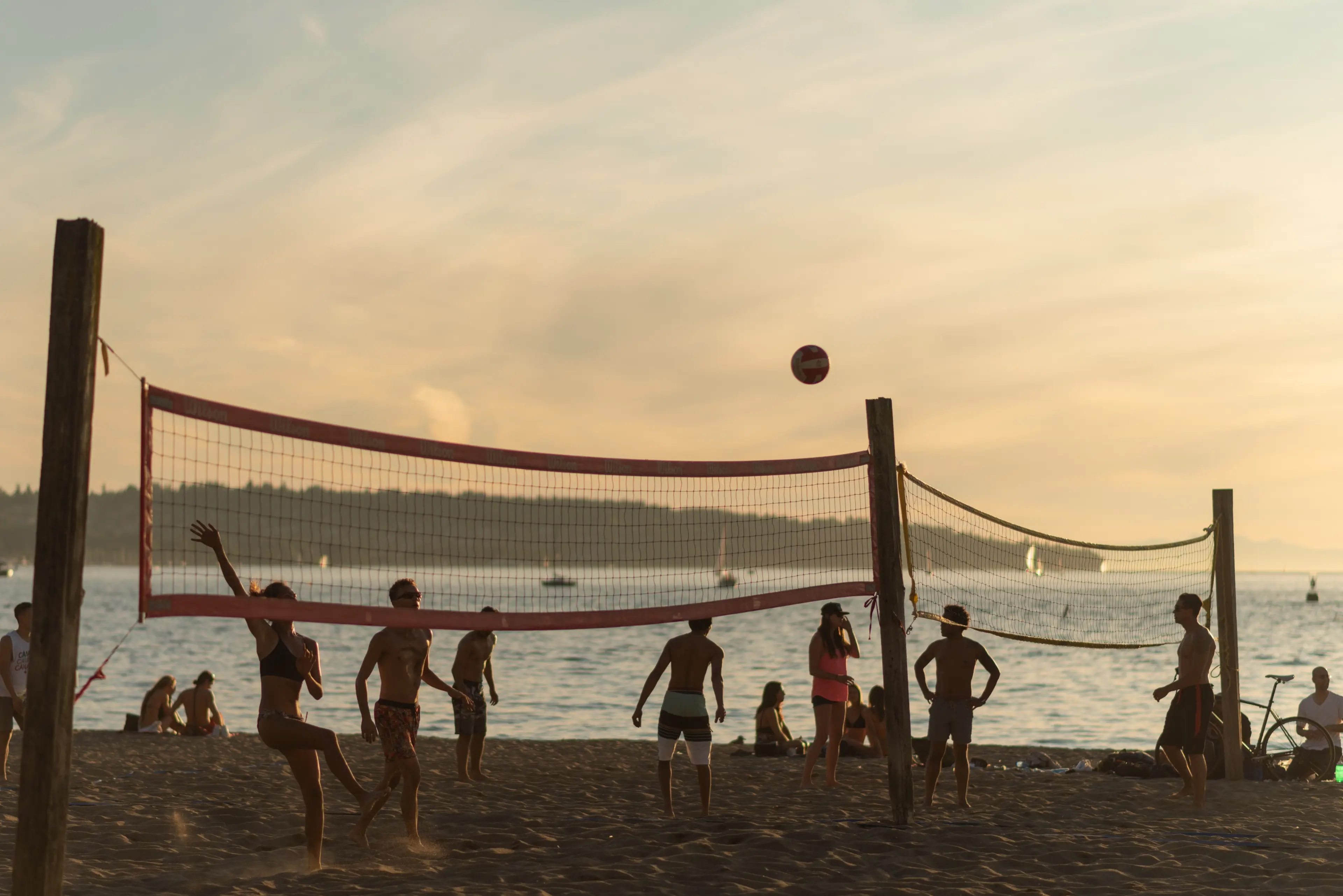Group of people playing beach volleyball near the ocean at sunset with a volleyball in mid-air.