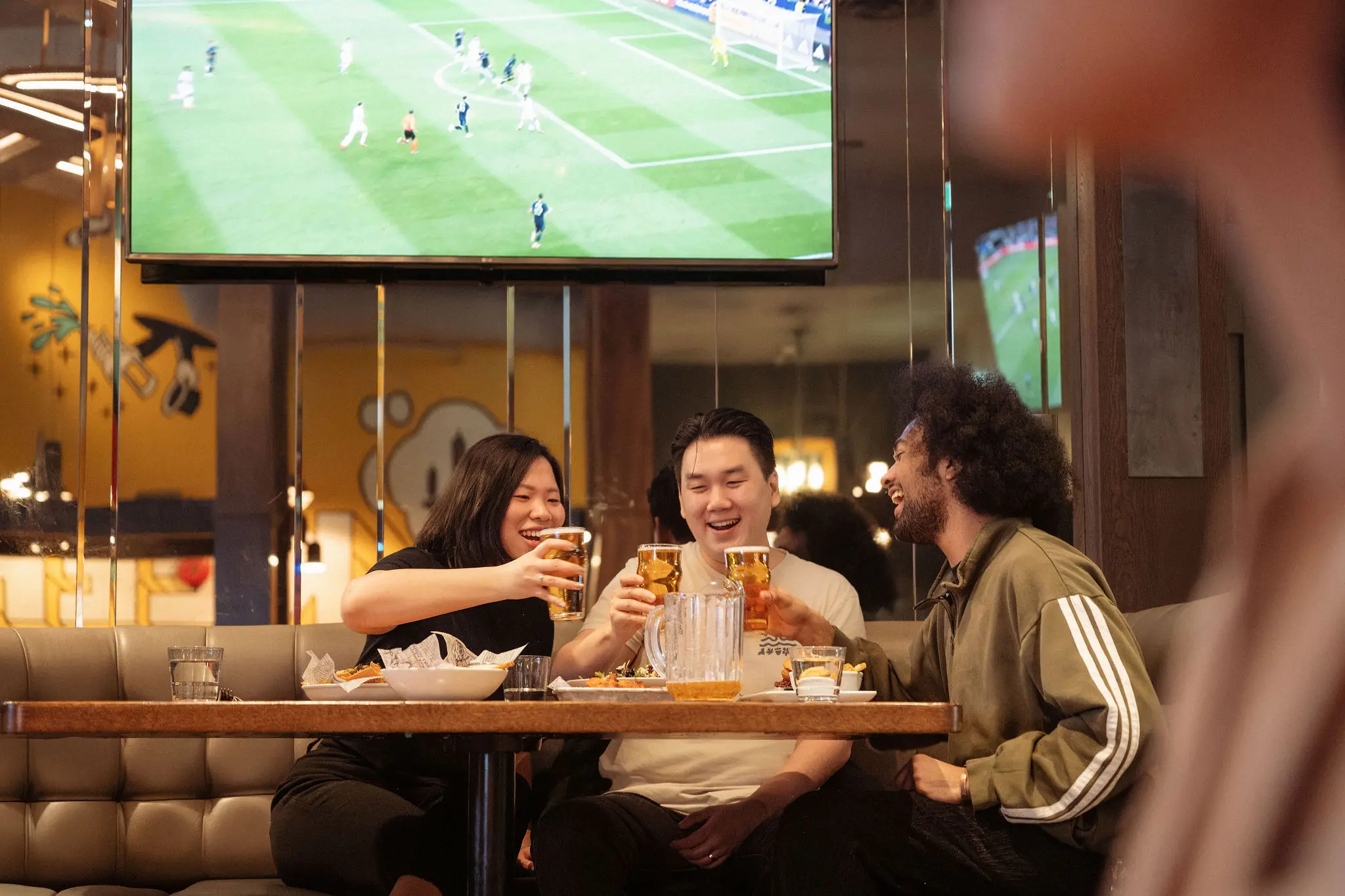 Three people sitting at a table in a bar, clinking glasses of beer, with a soccer game on a TV screen above them.