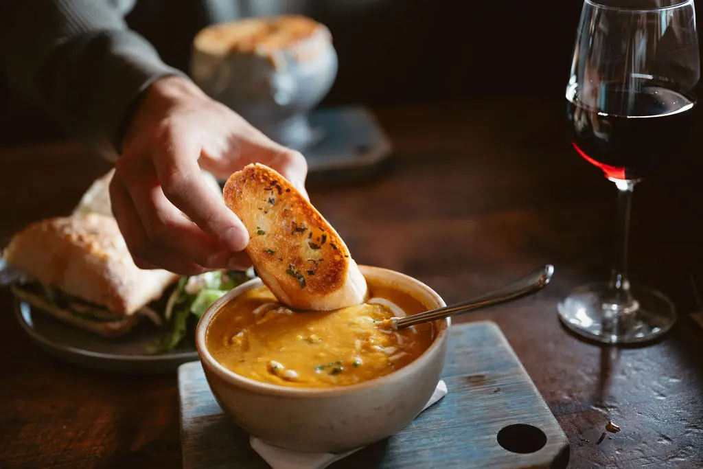 A hand dipping a piece of toasted baguette into a bowl of soup at the Burgoo restaurant in Vancouver.