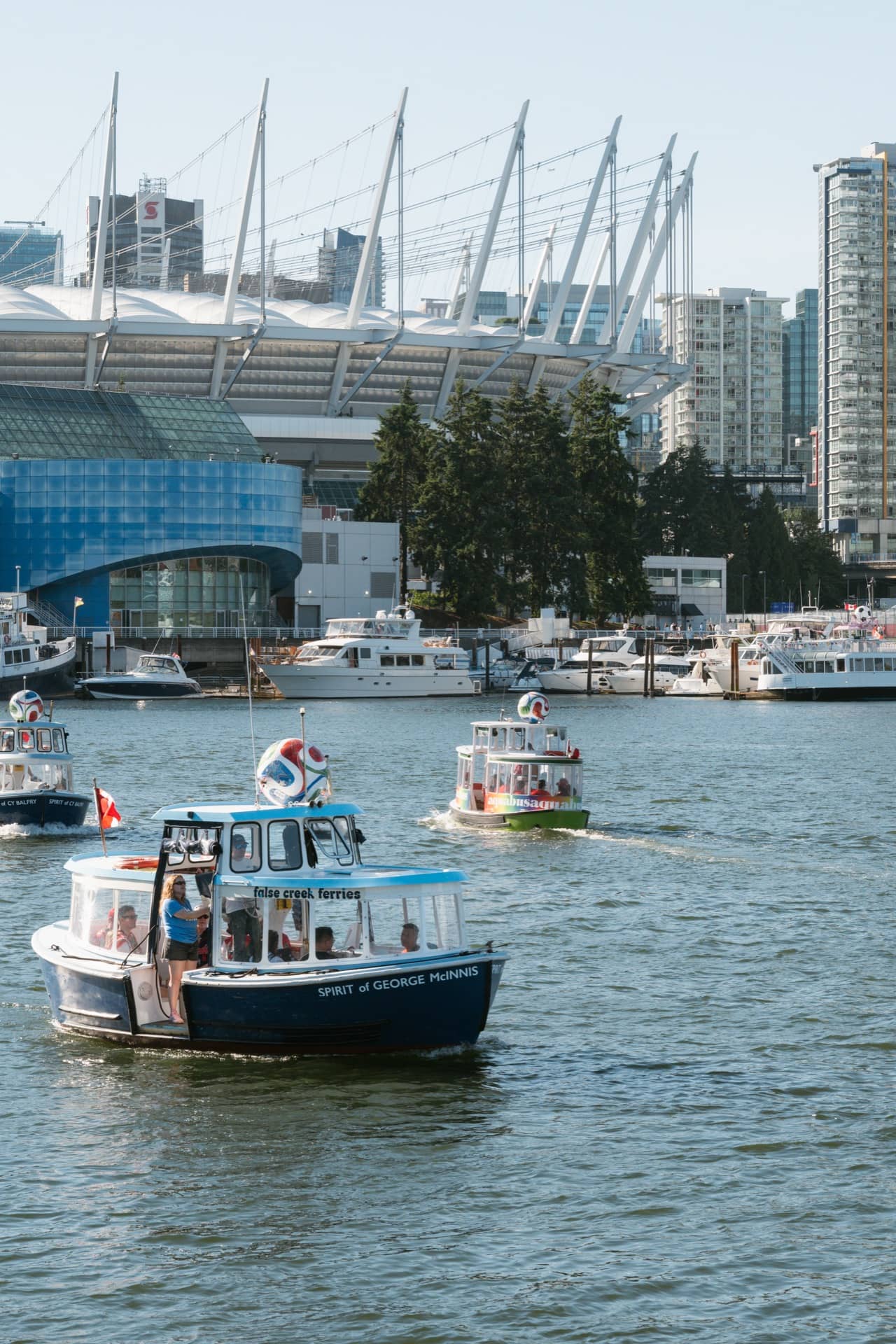 A small colorful Aquabus ferry with passengers crosses the water in a city harbor, surrounded by modern high-rise buildings under a clear sky, capturing the lively spirit ahead of the FIFA Vancouver World Cup events.