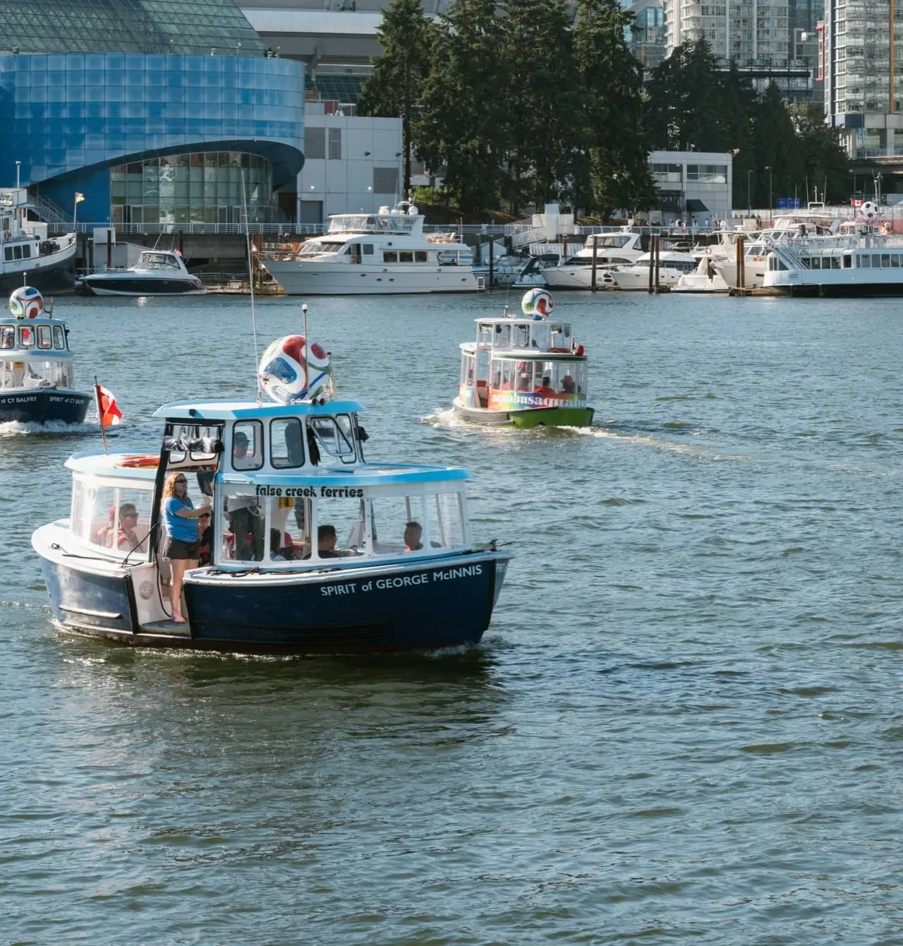 A small colorful Aquabus ferry with passengers crosses the water in a city harbor, surrounded by modern high-rise buildings under a clear sky, capturing the lively spirit ahead of the FIFA Vancouver World Cup events.