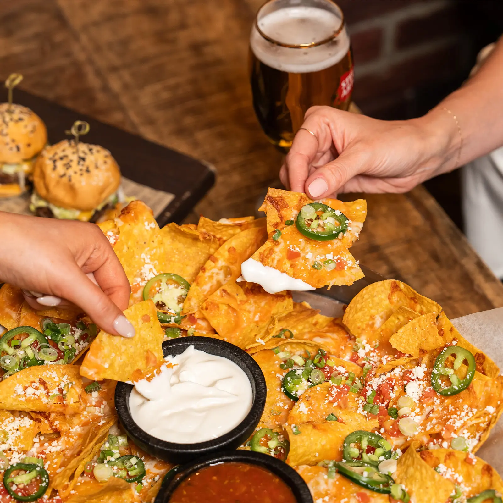 Two hands dipping jalapeño nachos with cheese and sour cream into sauces on a wooden table with sliders and a beer in the background.