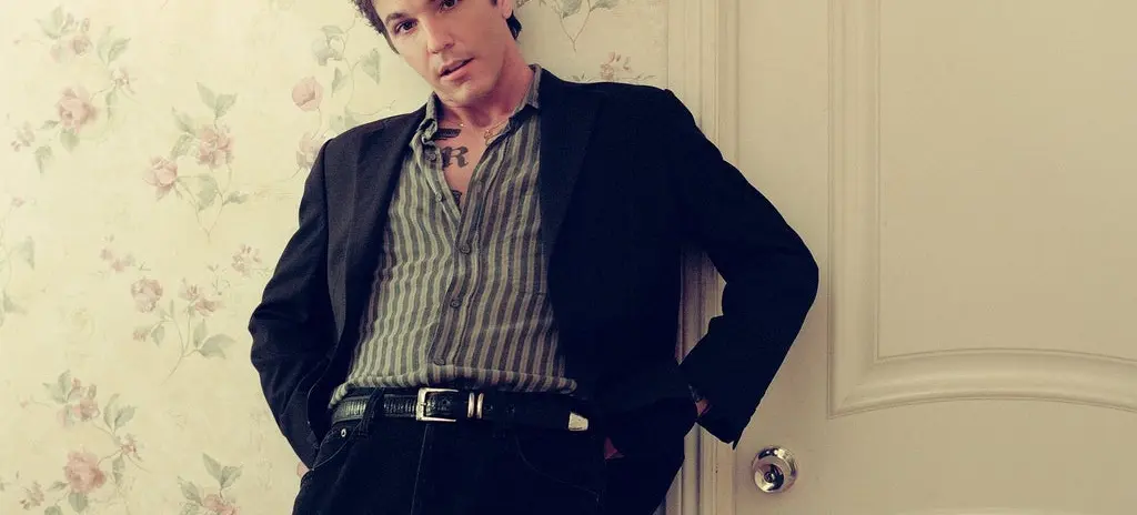 Person with short dark hair wearing a striped shirt and black jacket leaning against a floral wallpapered wall and a white door.
