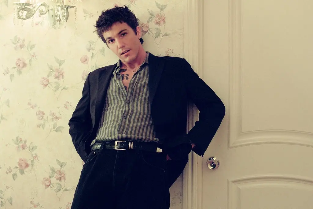 Person with short dark hair wearing a striped shirt and black jacket leaning against a floral wallpapered wall and a white door.