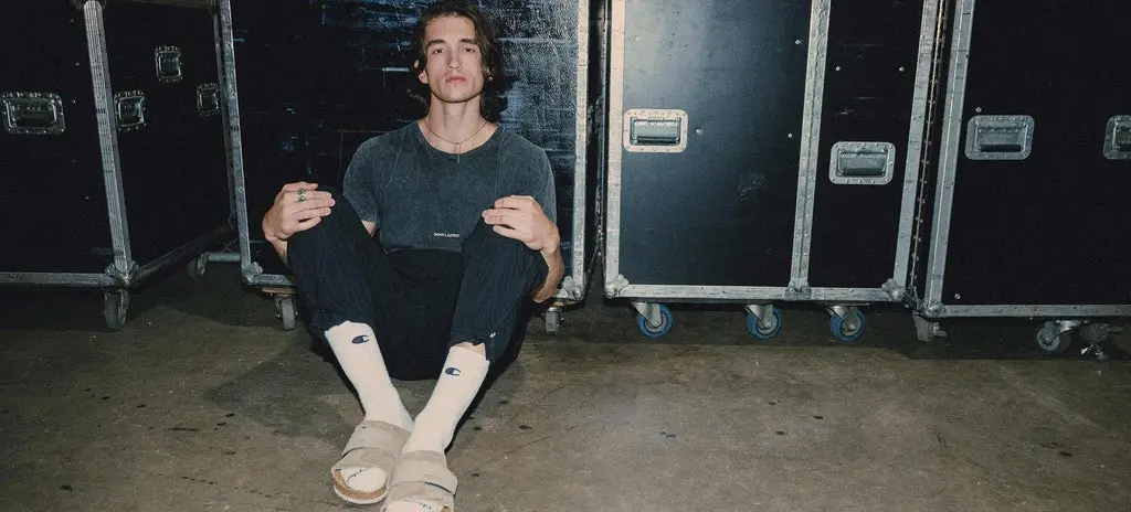 Person sitting on the floor with knees up, wearing a dark shirt, black pants, white socks, and beige sandals, against a backdrop of black equipment cases.