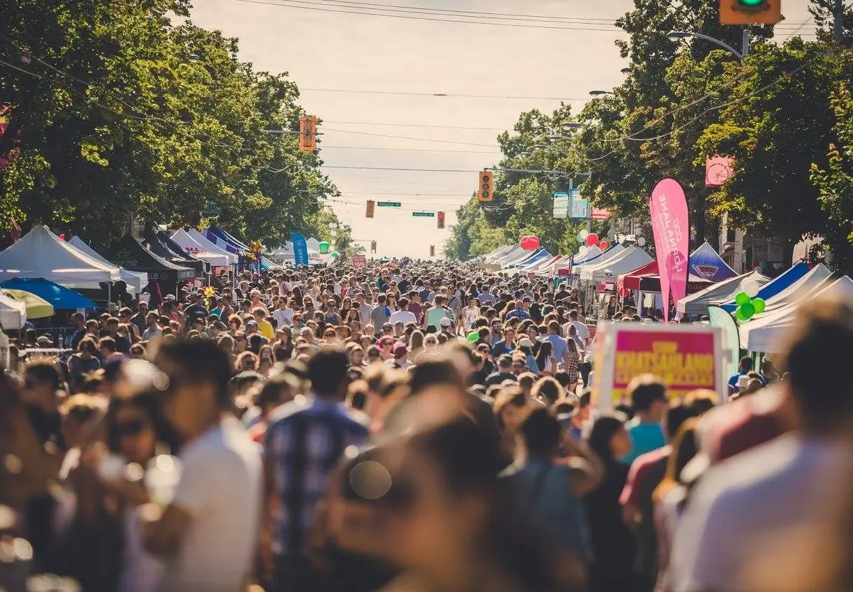 Crowded outdoor street market with many people walking between rows of vendor tents and trees lining the street.