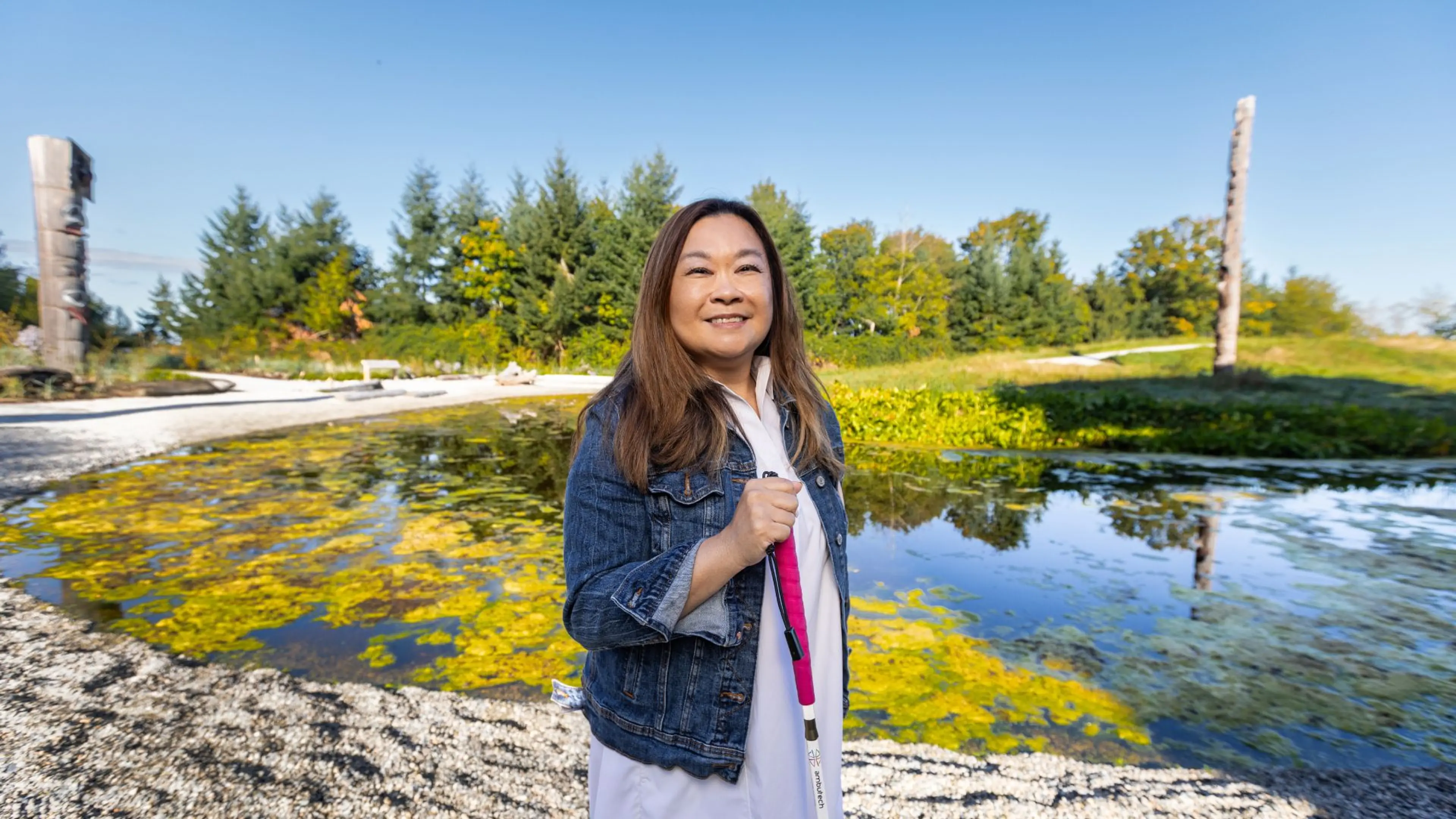 A smiling woman holding a white mobility cane stands by a tranquil pond and carved wooden poles at a lush outdoor park.