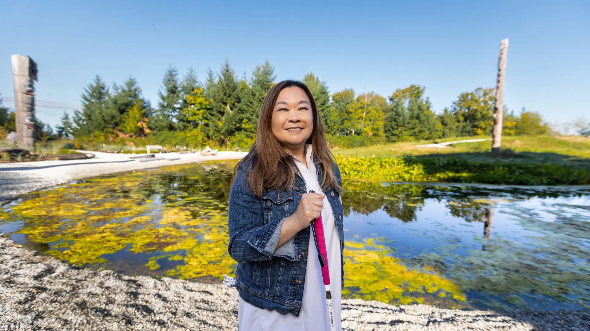 A smiling woman holding a white mobility cane stands by a tranquil pond and carved wooden poles at a lush outdoor park.