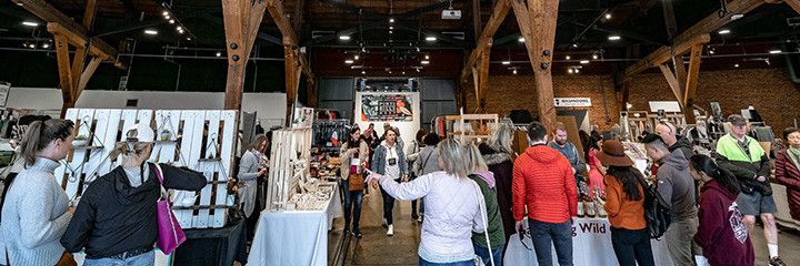 Indoor market with people browsing various stalls displaying crafts and goods under wooden beams.