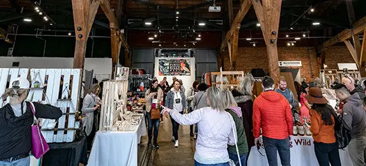 Indoor market with people browsing various stalls displaying crafts and goods under wooden beams.