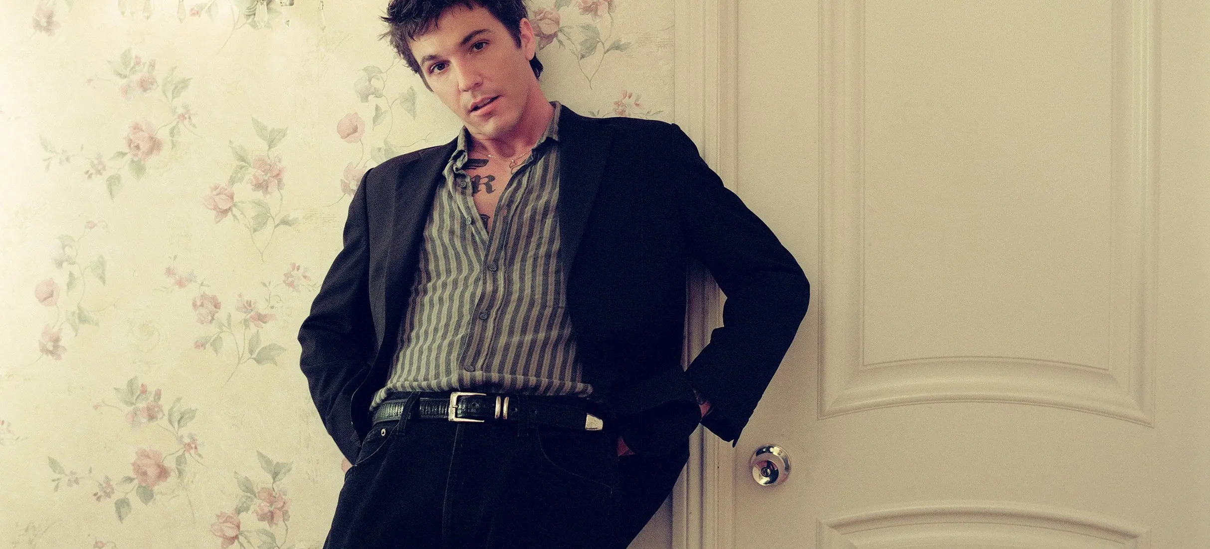 Person leaning against a door with hands in pockets, wearing a striped shirt and black jacket in a floral wallpapered room.
