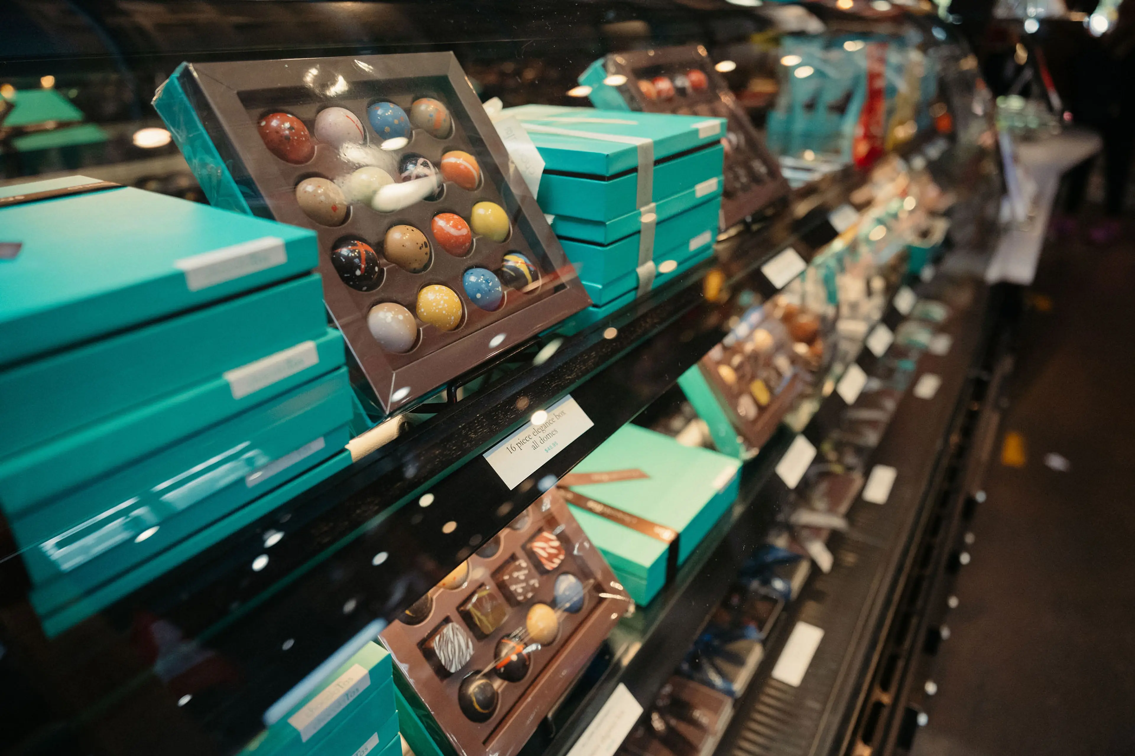 Display case with assorted colorful chocolates and teal gift boxes in a store.