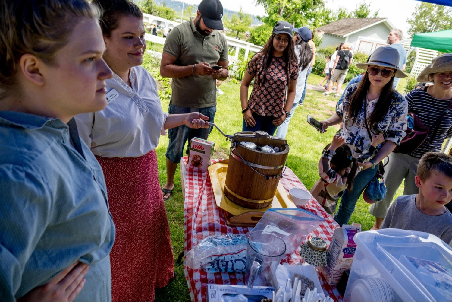 People gathered around a table with a red and white checkered tablecloth outdoors, with an ice cream maker and supplies on the table.