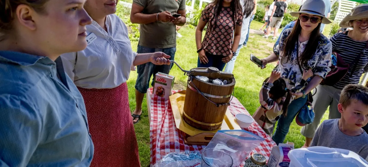 People gathered around a table with a red and white checkered tablecloth outdoors, with an ice cream maker and supplies on the table.