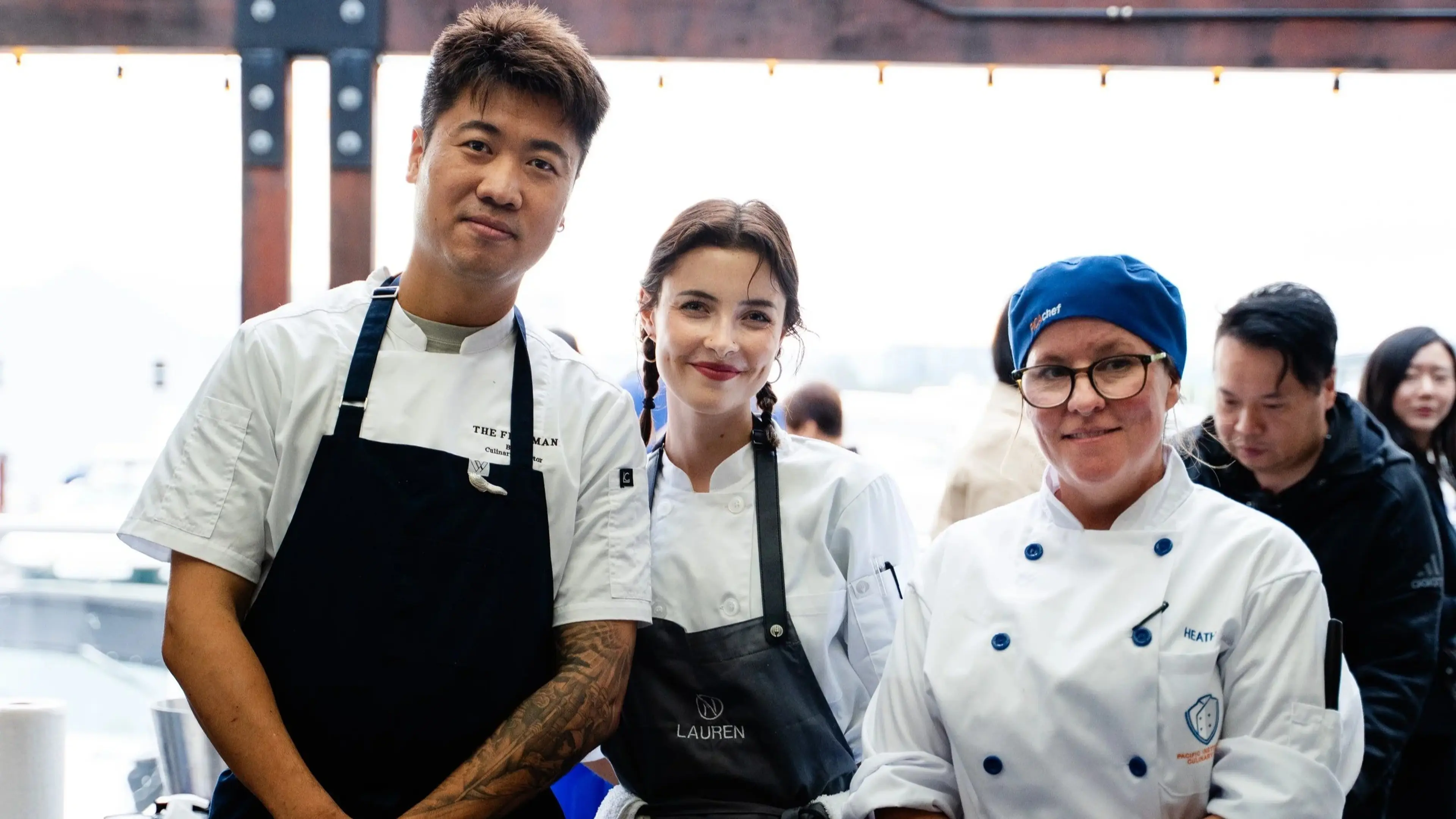 Three chefs in white uniforms and aprons posing together, one wearing a blue chef hat and glasses.