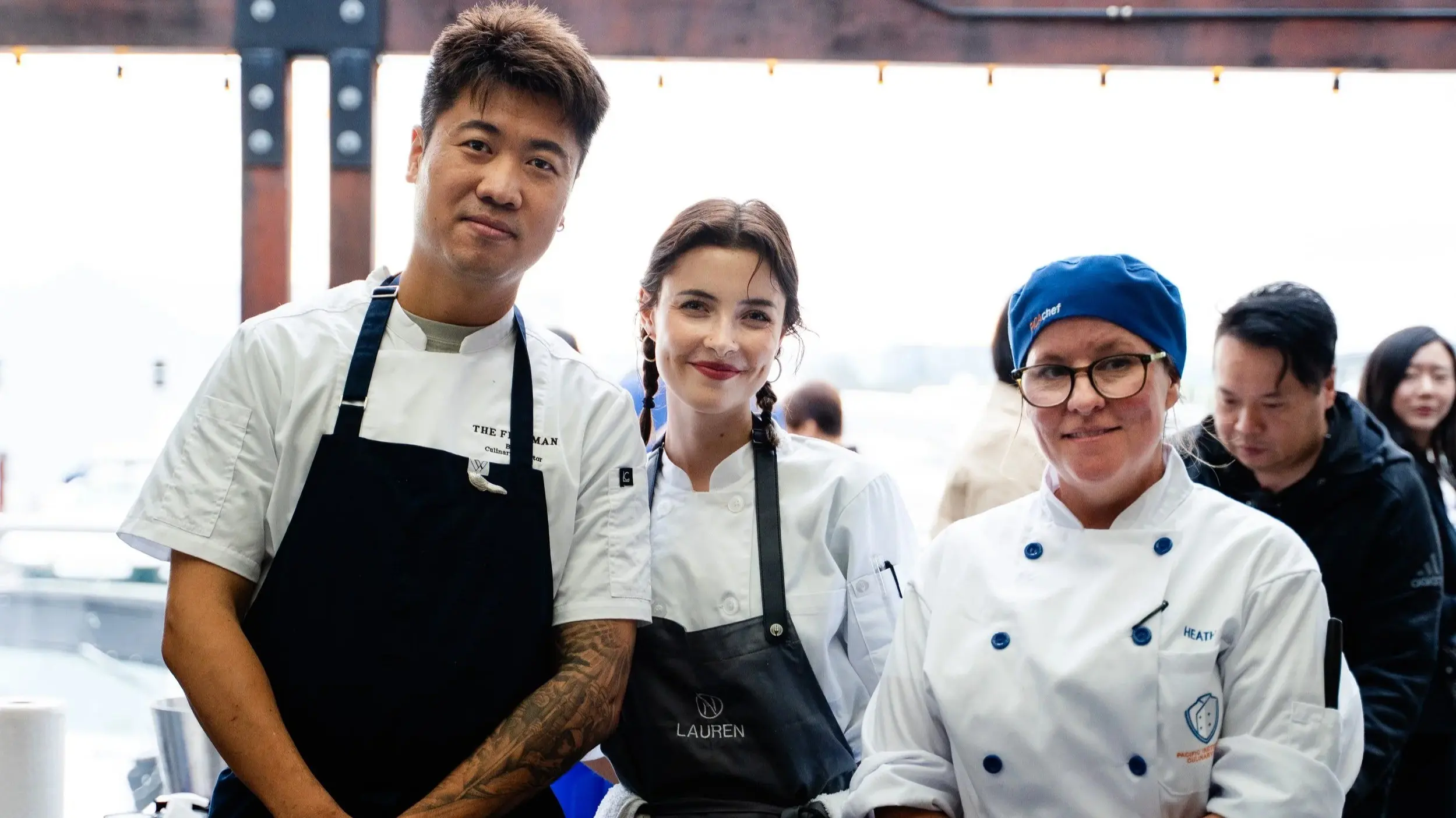 Three chefs in white uniforms and aprons posing together, one wearing a blue chef hat and glasses.