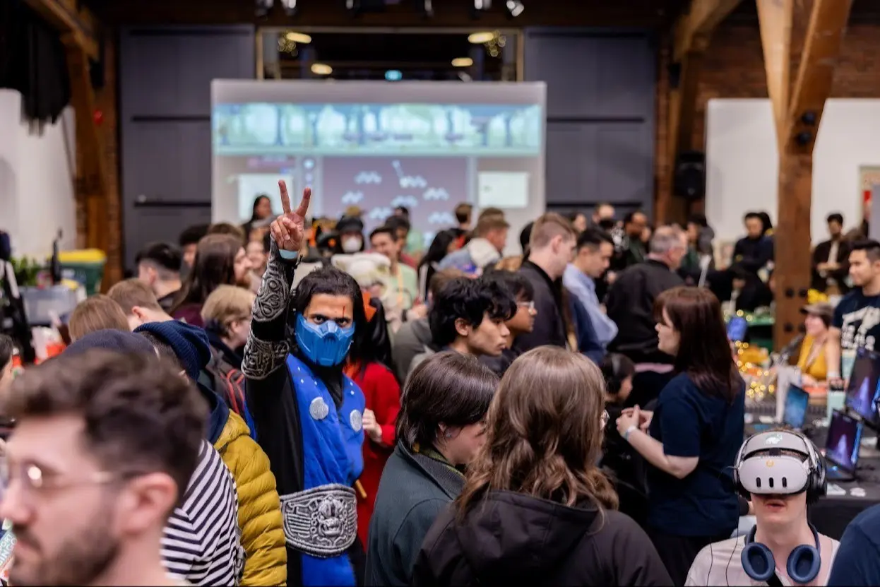 Crowded indoor event with a person in blue costume and mask making a peace sign, and another person wearing a VR headset.