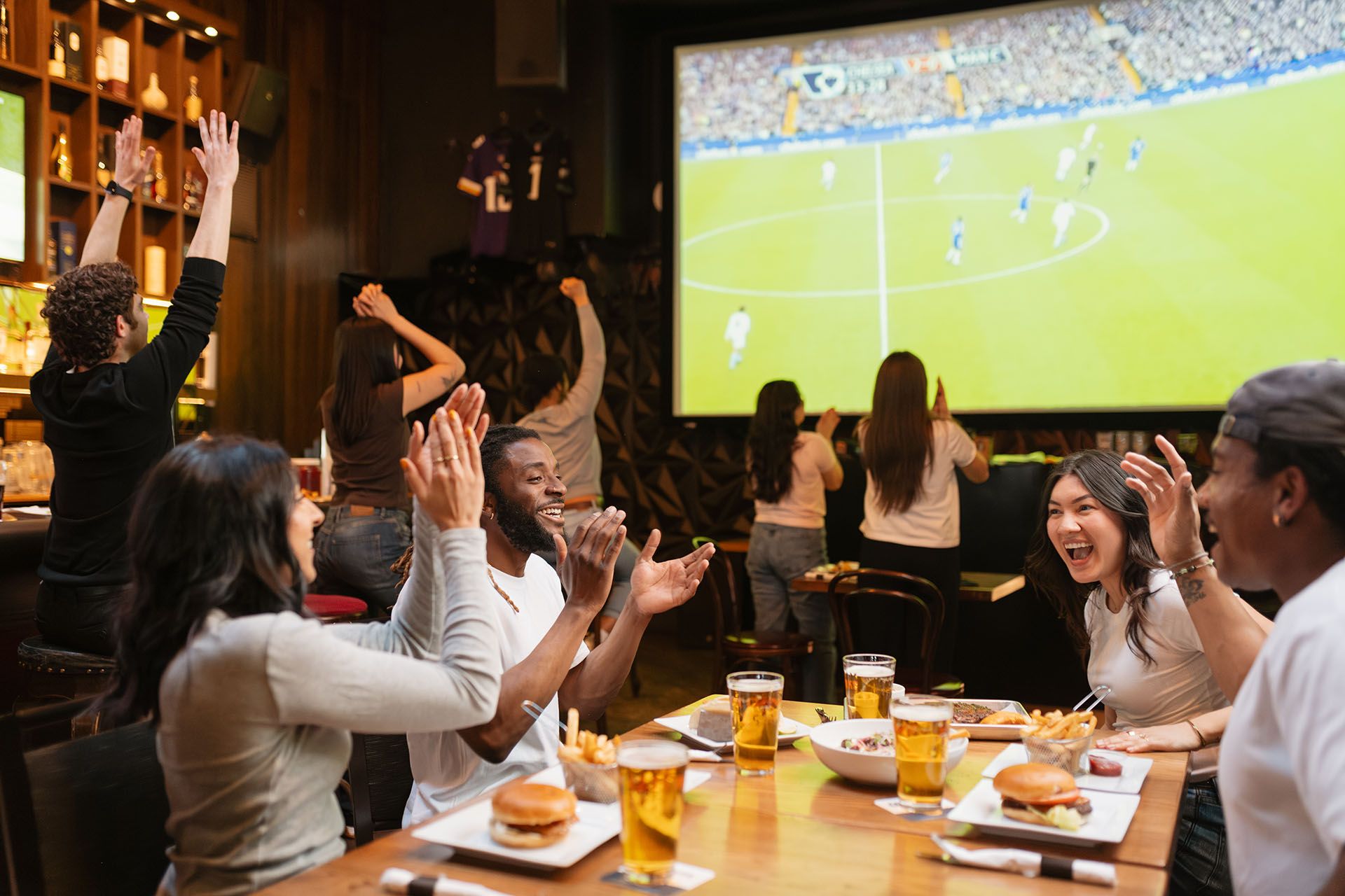 Group of diverse friends cheering and clapping while watching a soccer game on a large screen in a bar with food and drinks on the table.
