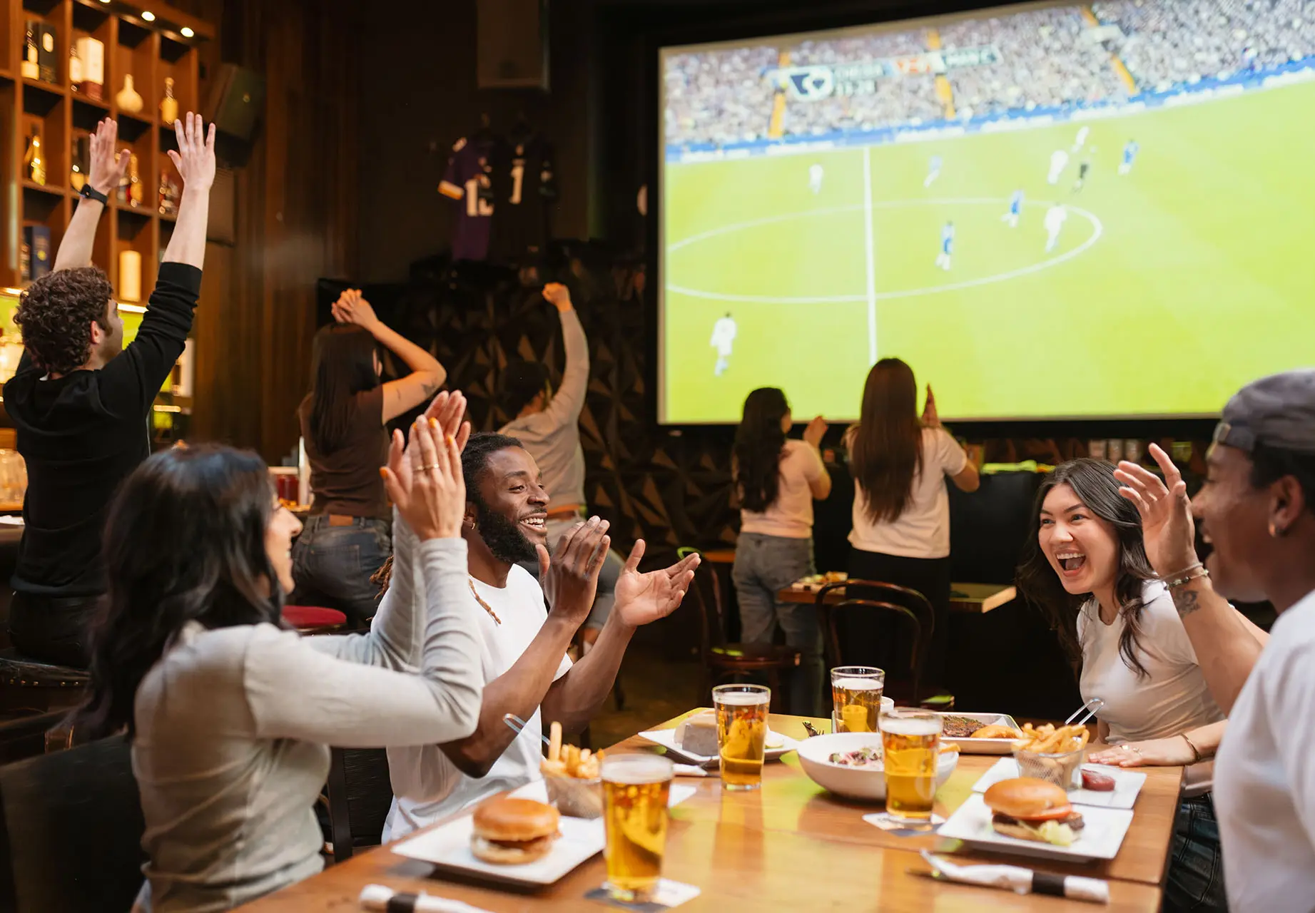 Group of diverse friends cheering and clapping while watching a soccer game on a large screen in a bar with food and drinks on the table.