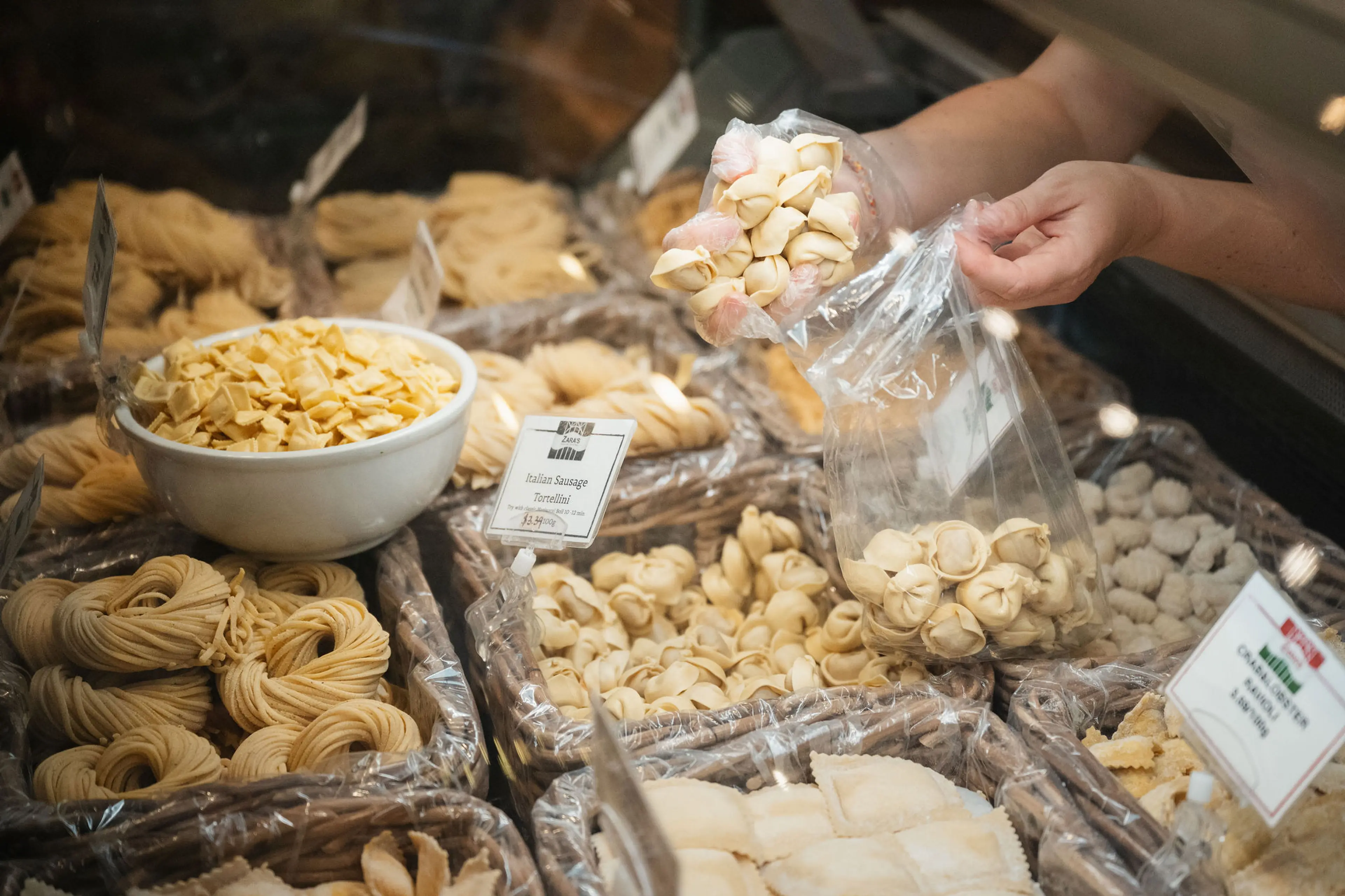 Person scooping fresh tortellini pasta into a plastic bag at a deli counter with various types of pasta displayed.