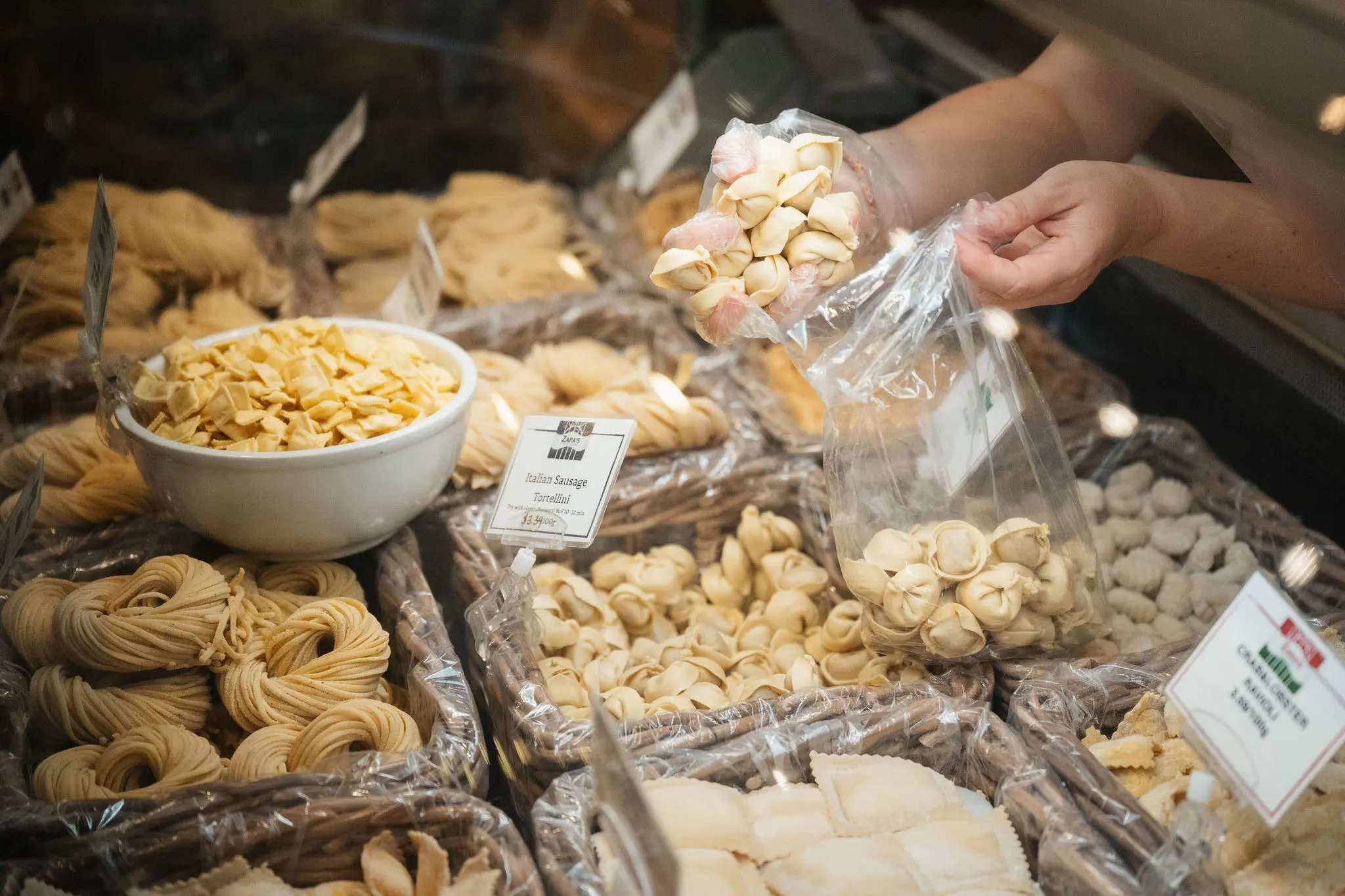 Person scooping fresh tortellini pasta into a plastic bag at a deli counter with various types of pasta displayed.