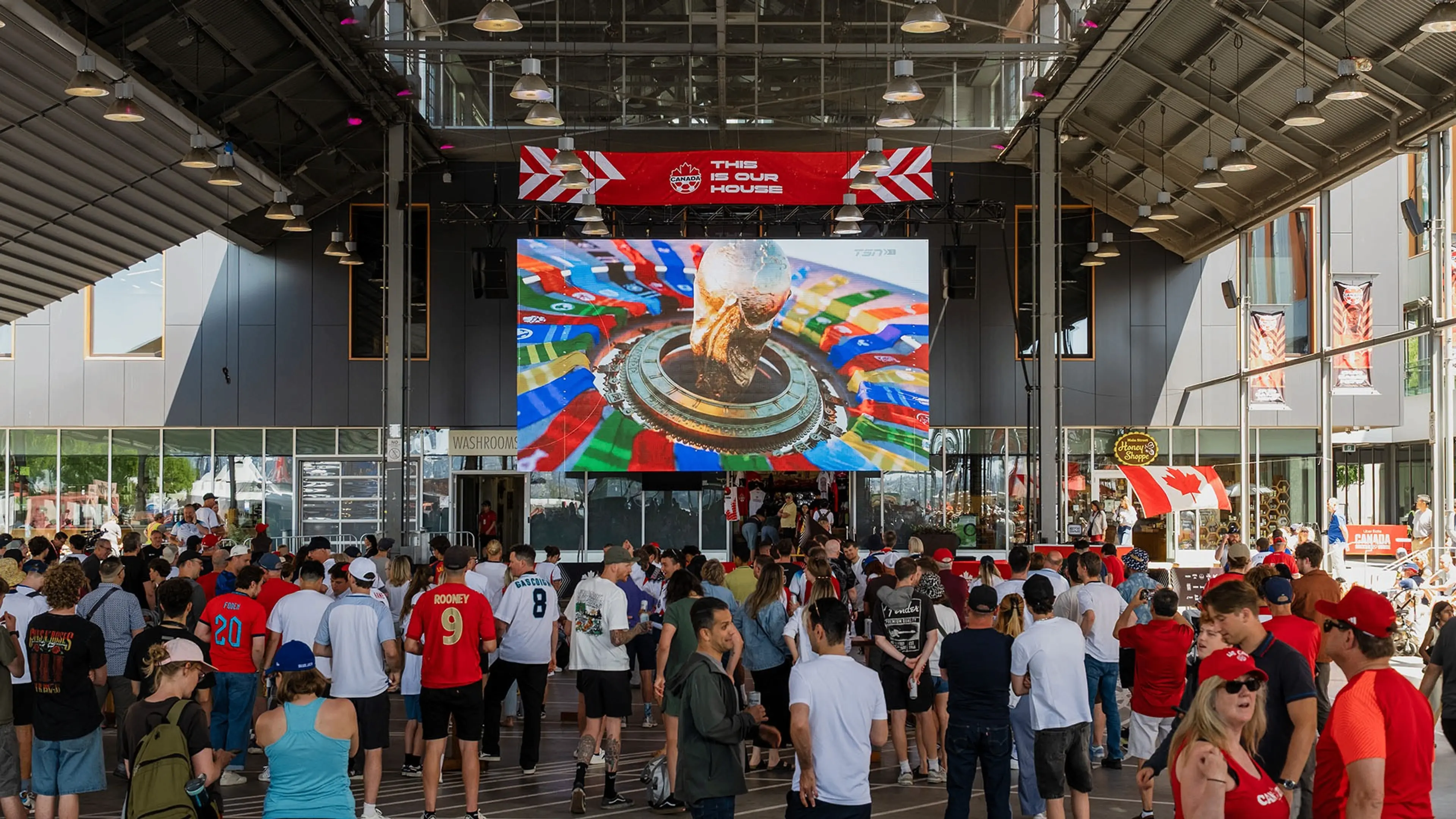 Crowd of people watching a large screen showing a World Cup trophy inside a covered outdoor area with Canadian flags.