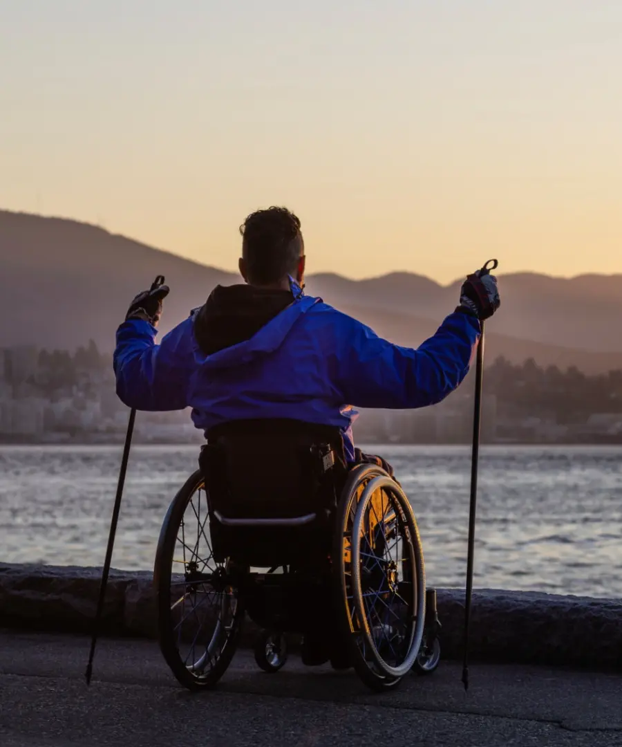 A person in a manual wheelchair holding trekking poles looks out over the ocean and mountains at sunrise from the paved Stanley Park Seawall.