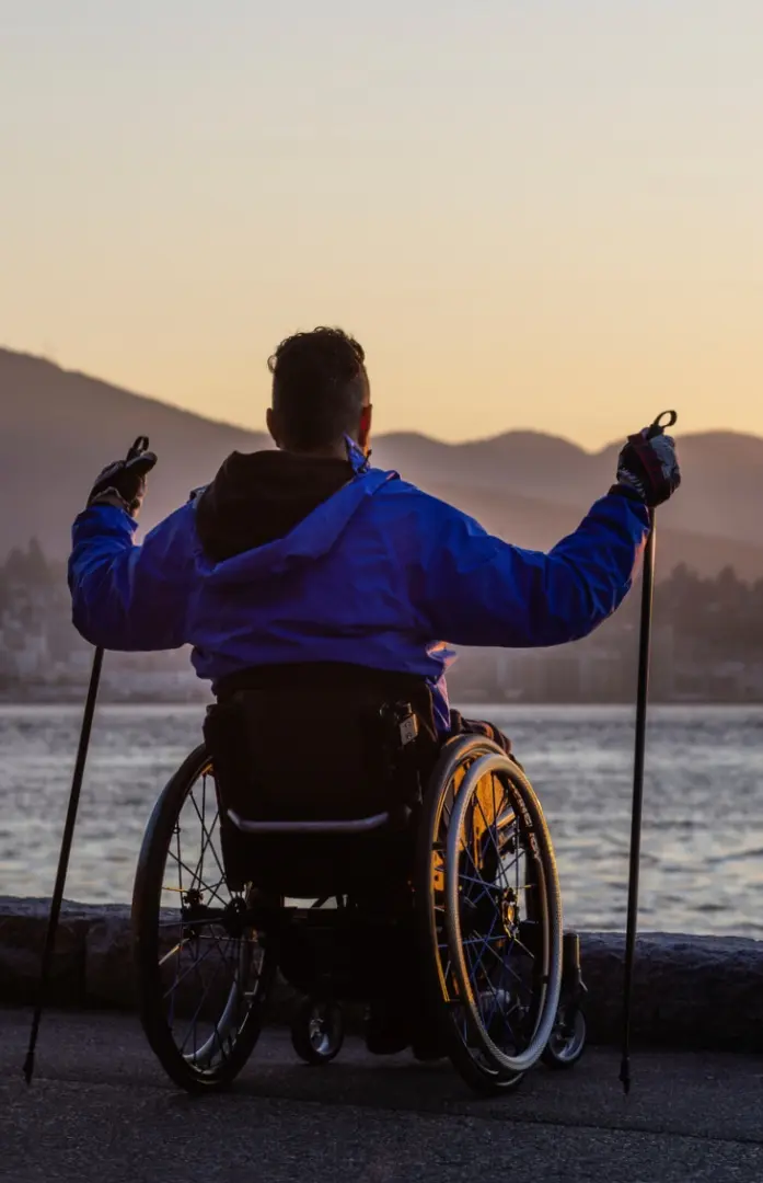 A person in a manual wheelchair holding trekking poles looks out over the ocean and mountains at sunrise from the paved Stanley Park Seawall.