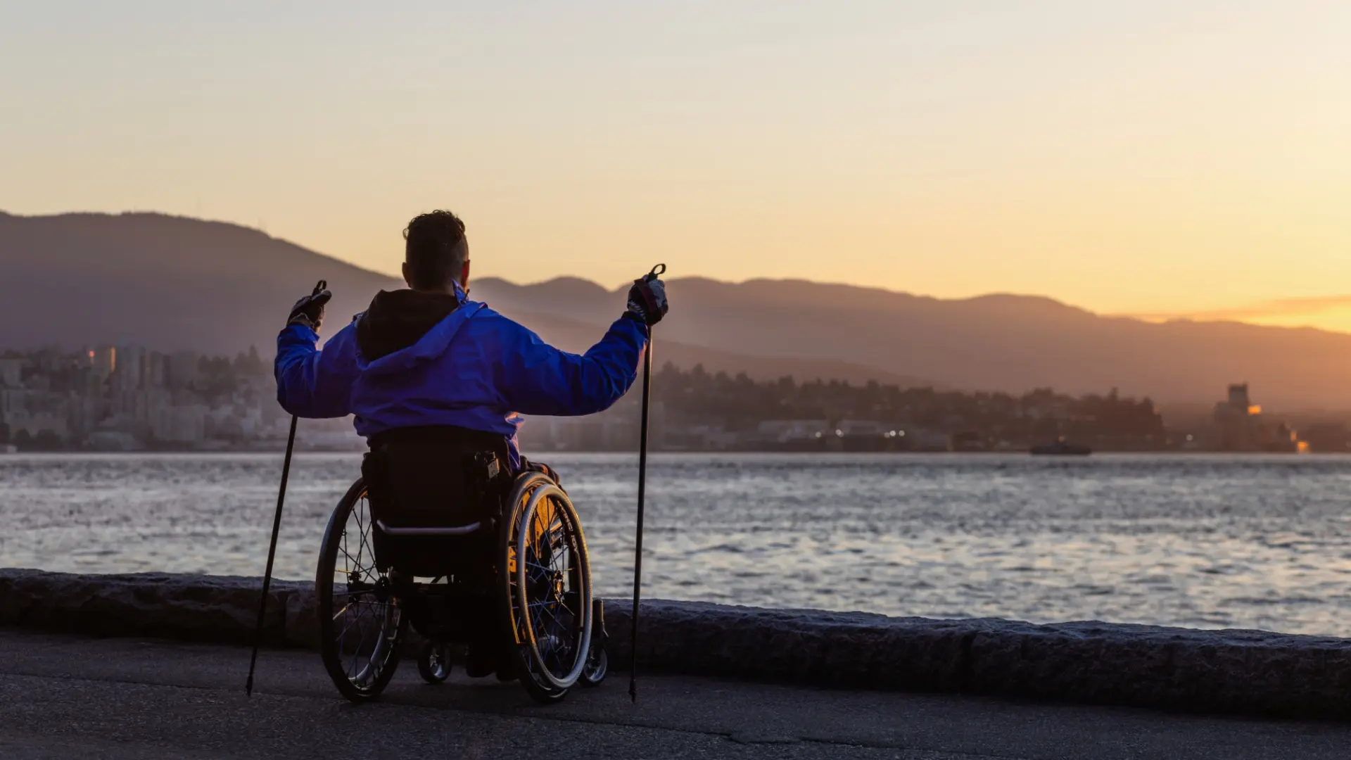 A person in a manual wheelchair holding trekking poles looks out over the ocean and mountains at sunrise from the paved Stanley Park Seawall.