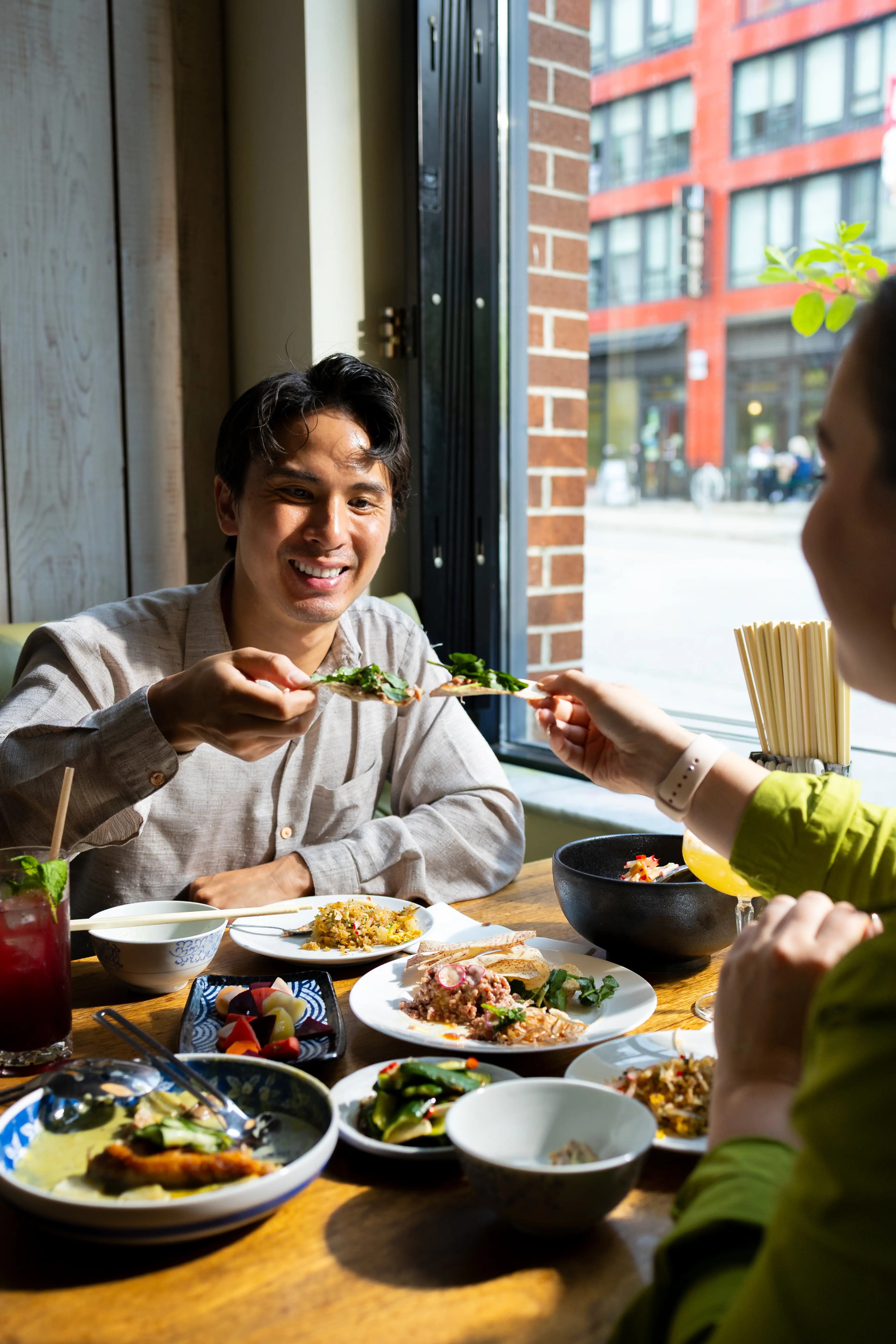 Two people sitting at a table by a window sharing food and smiling, with various dishes and drinks on the table.
