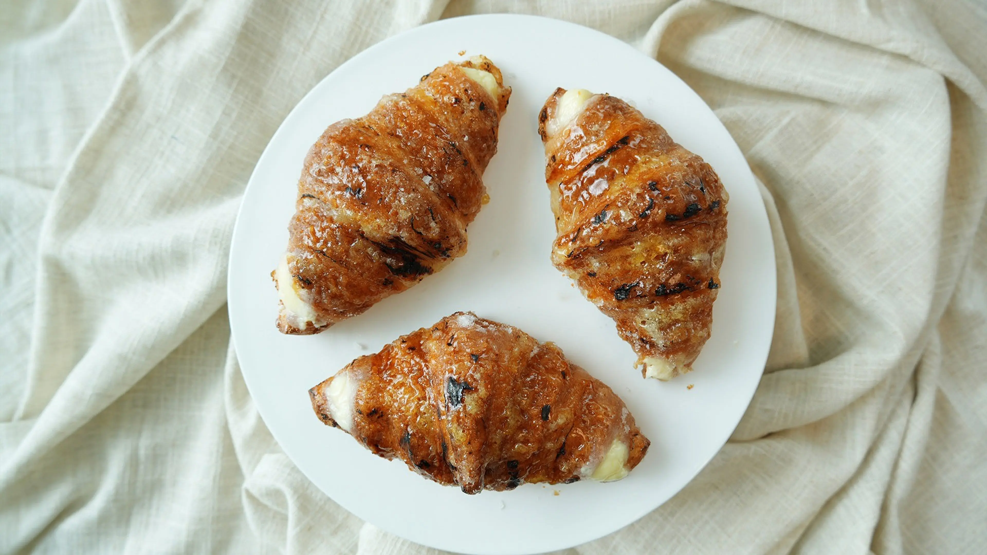 Three glazed croissants with cream filling on a white plate over a beige cloth.