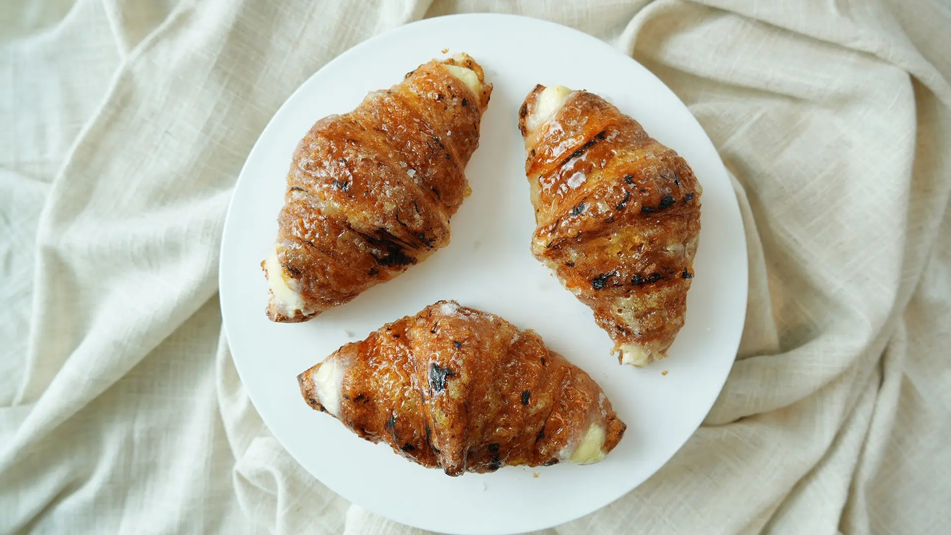 Three glazed croissants with cream filling on a white plate over a beige cloth.
