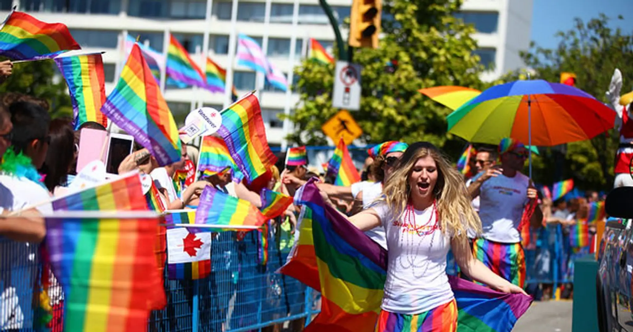 Person with long hair holding a rainbow flag and wearing rainbow skirt at a pride parade with crowd waving rainbow flags and a rainbow umbrella.
