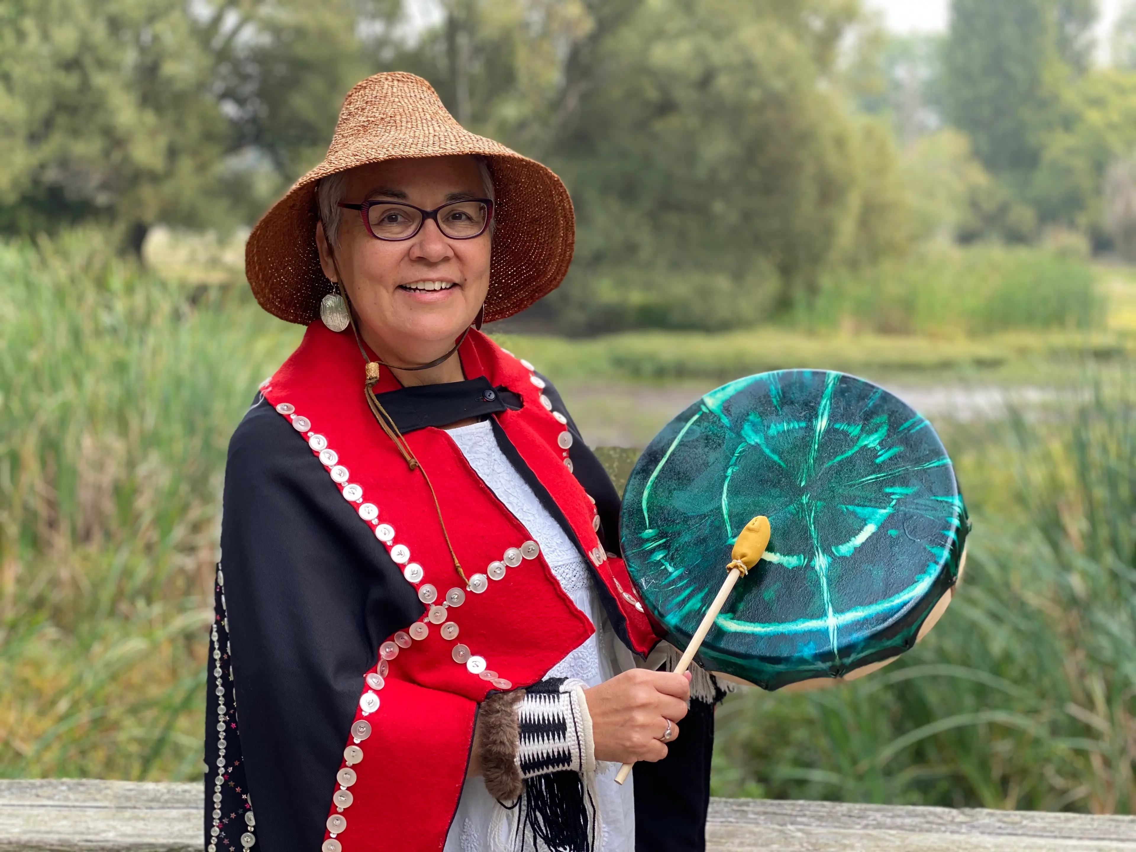 Smiling person wearing a woven hat and black garment decorated with silver buttons, holding a teal drum and drumstick outdoors.