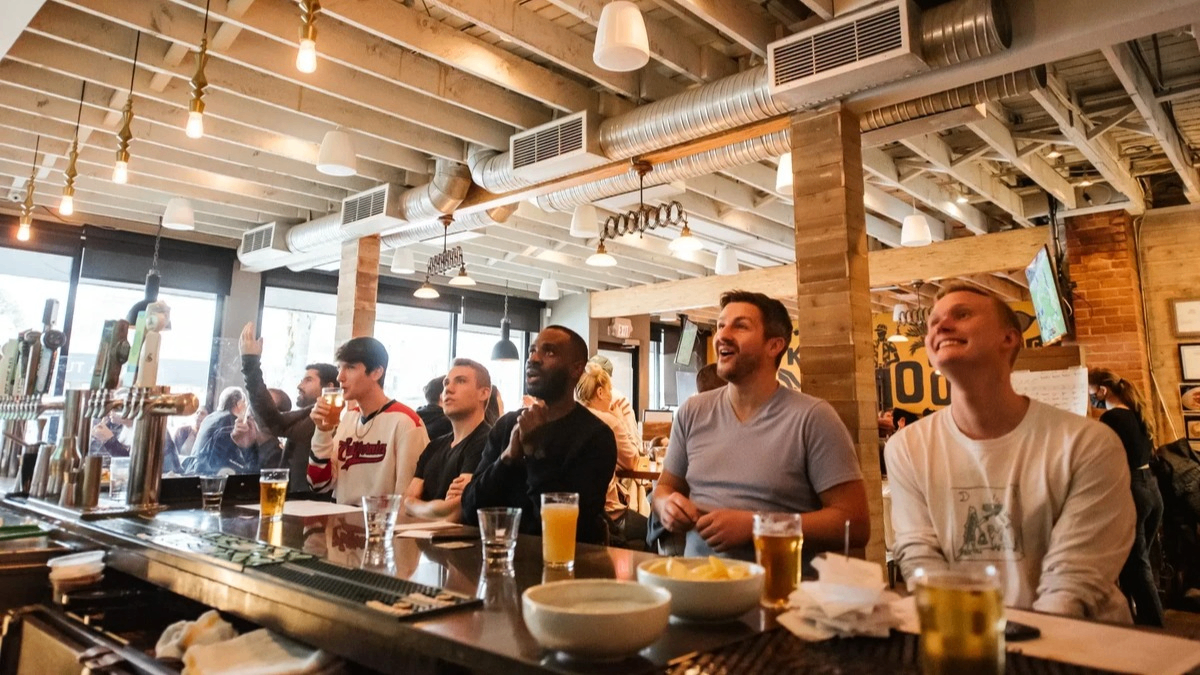 Six people sitting at a bar counter with drinks, watching something with focused and excited expressions.