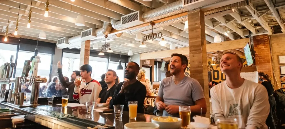 Six people sitting at a bar counter with drinks, watching something with focused and excited expressions.