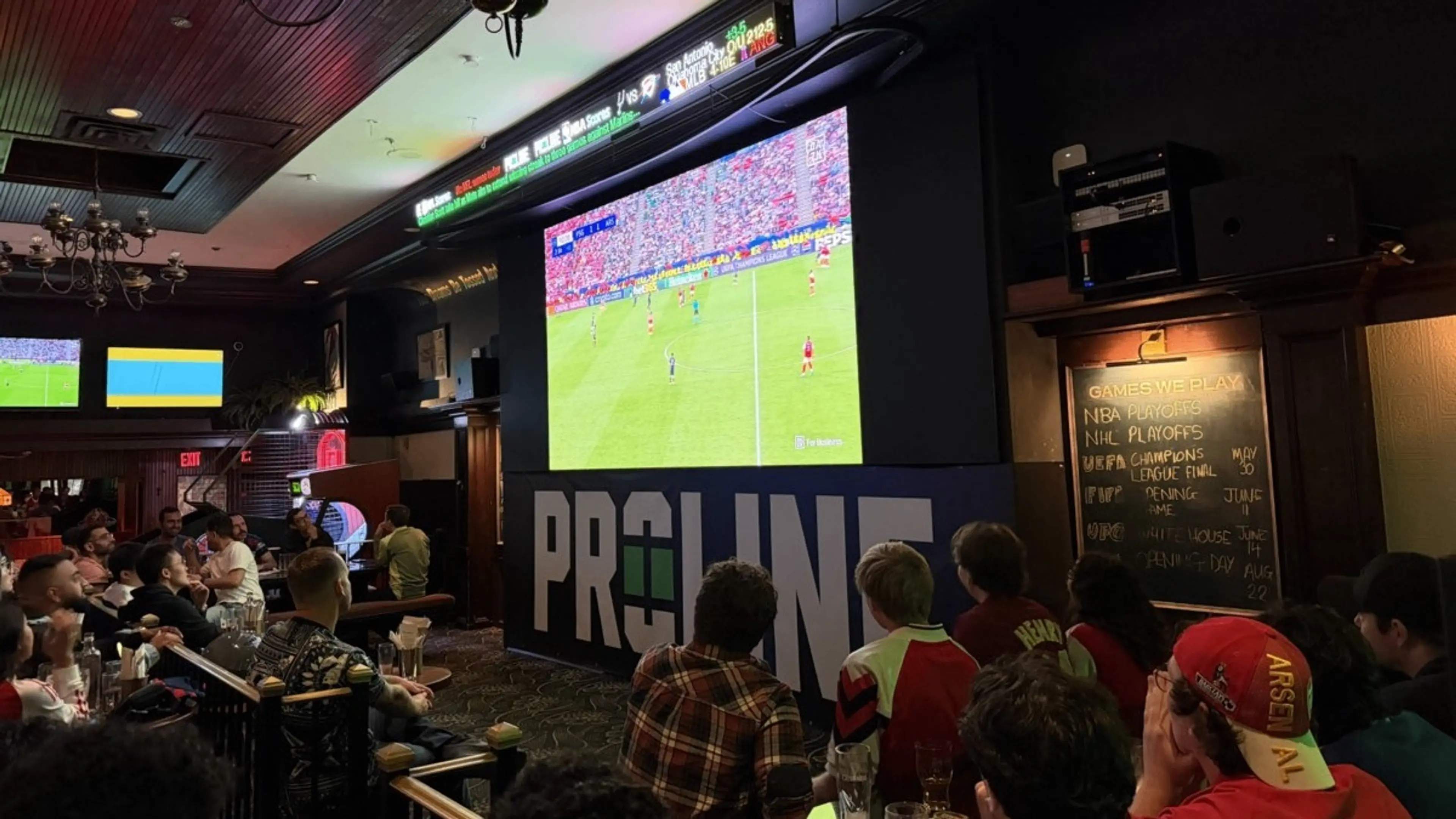 A group of people watching a soccer game on a large screen in a dimly lit sports bar with a scoreboard and a chalkboard listing games.