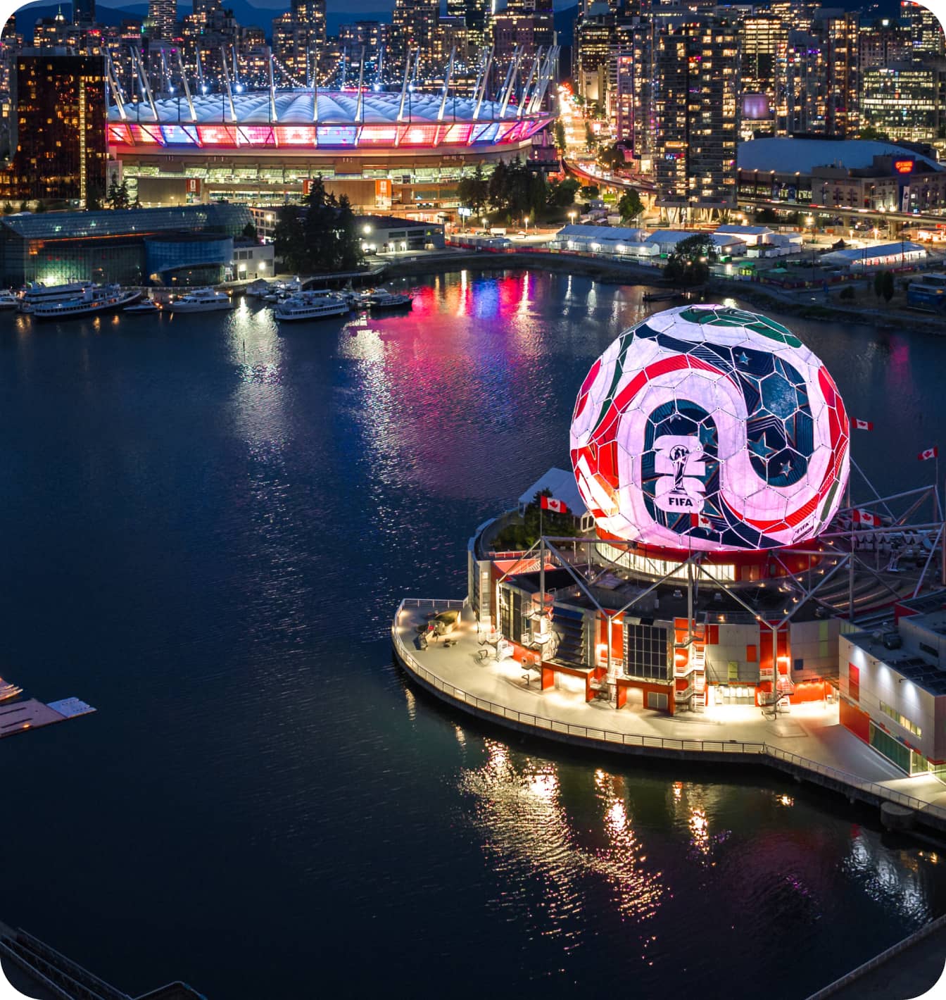 Night aerial view of a waterfront city with a large illuminated spherical structure displaying FIFA logo and a stadium lit in red and blue in the background.