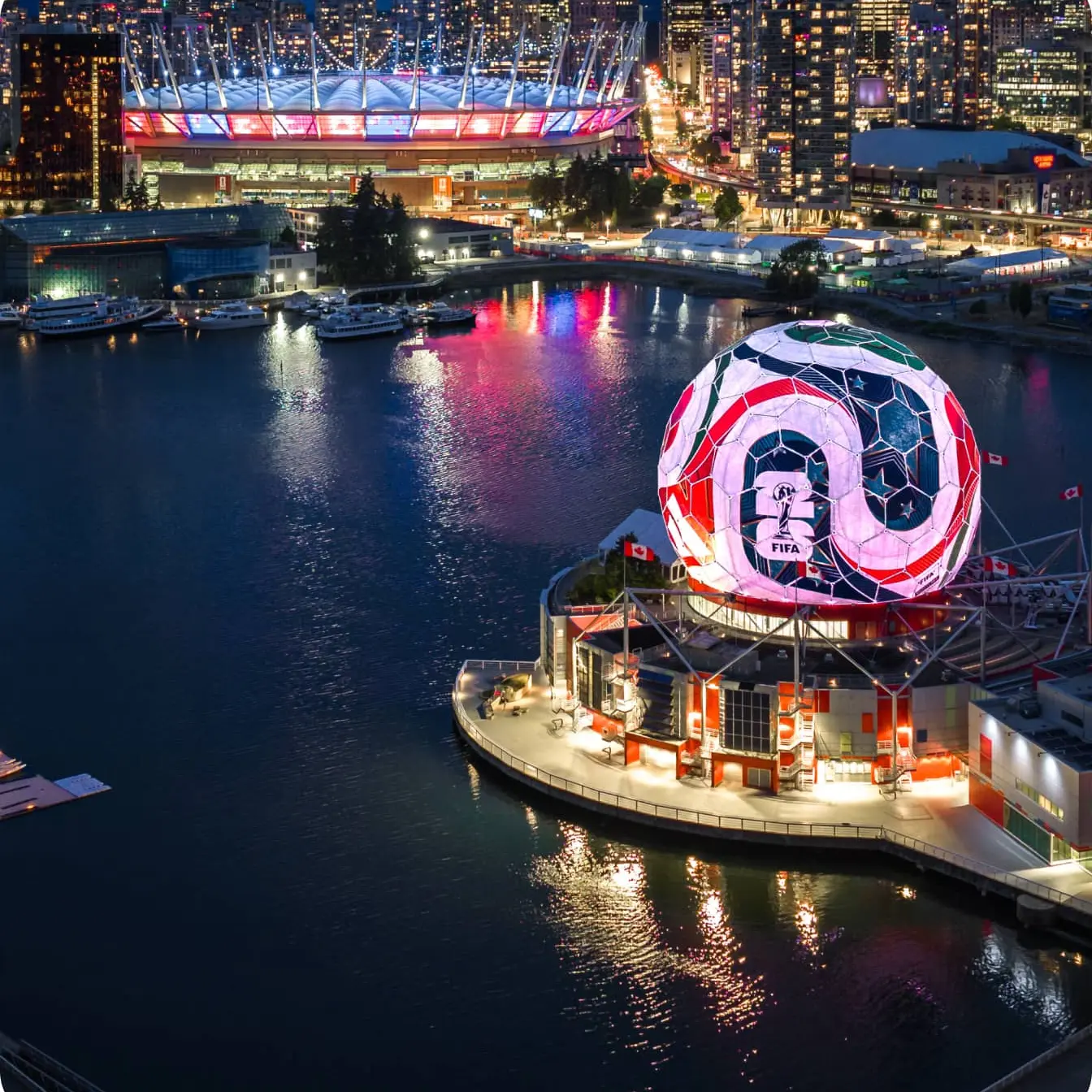 Night aerial view of a waterfront city with a large illuminated spherical structure displaying FIFA logo and a stadium lit in red and blue in the background.
