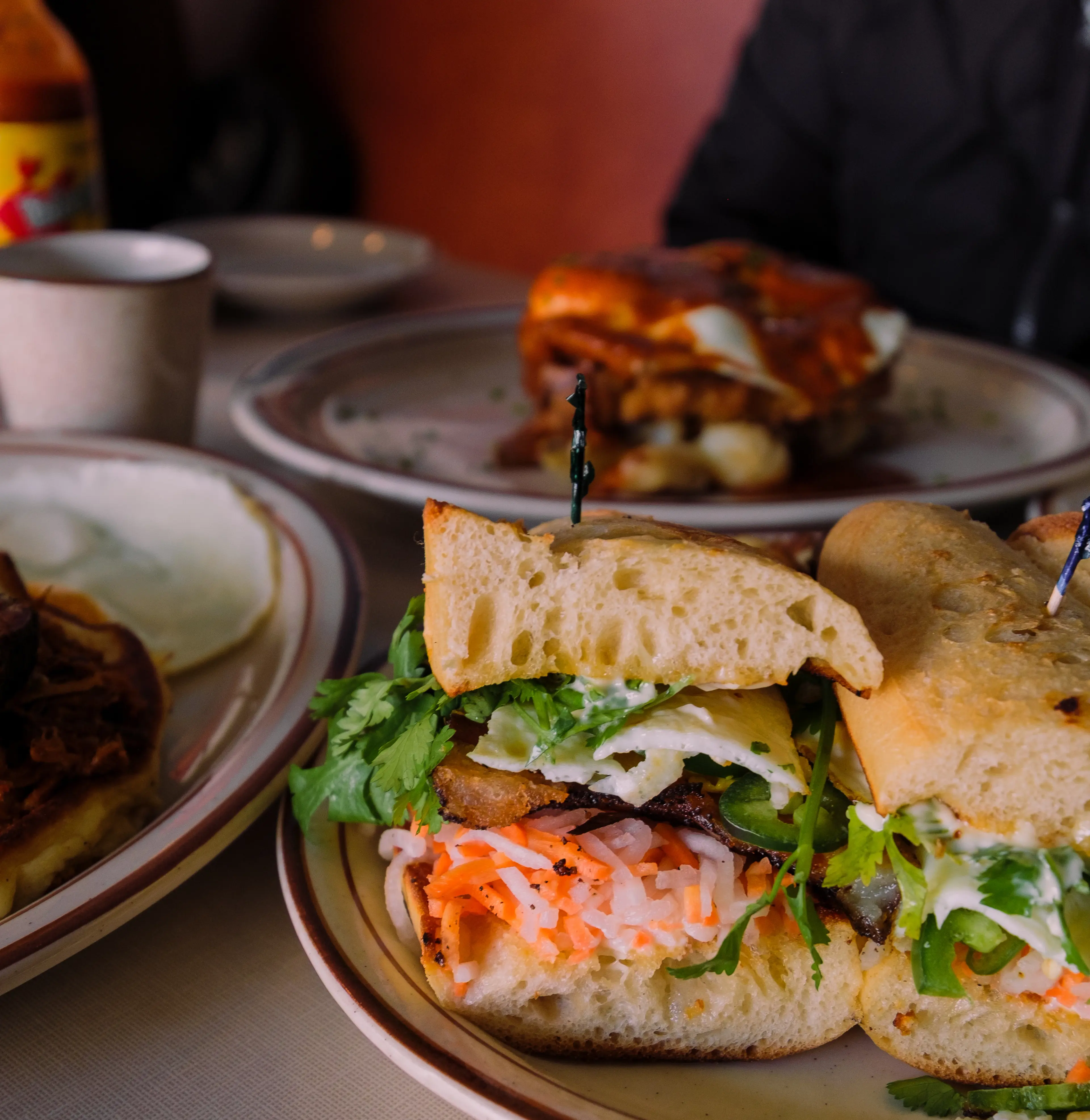 Close-up of a sandwich with pickled vegetables, greens, and meat on a plate, with other plates and a cup in the background.