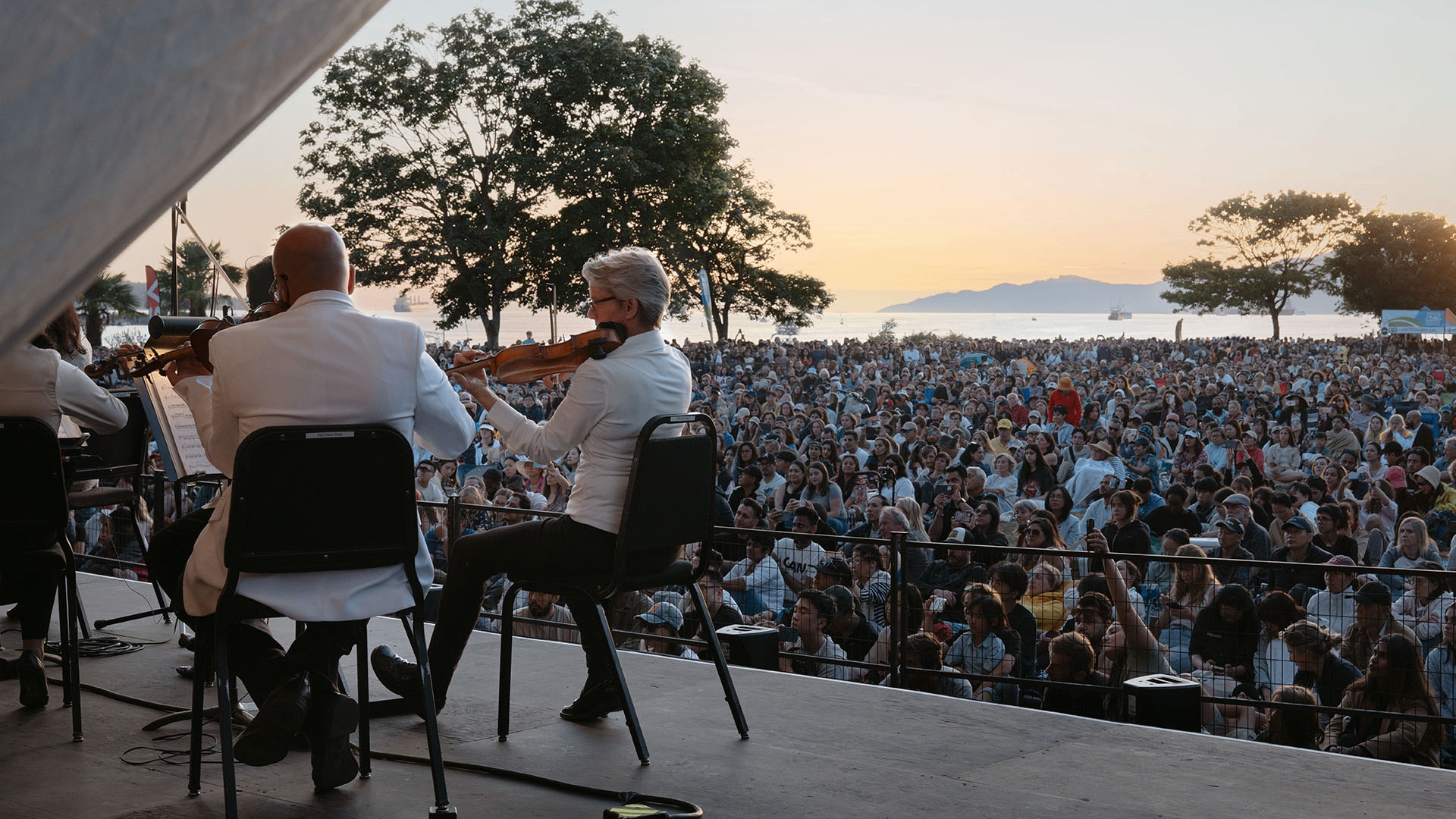 Musicians in white jackets playing violins on stage facing a large outdoor audience at sunset near water and trees.